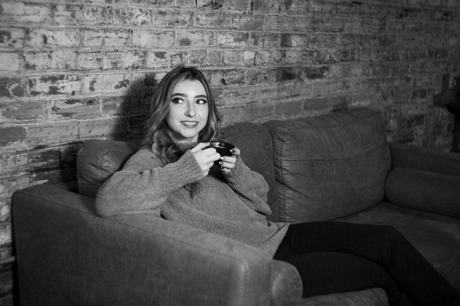 Black and white photo of a young woman relaxing on a gray sofa, holding a camera, smiling near a brick wall.