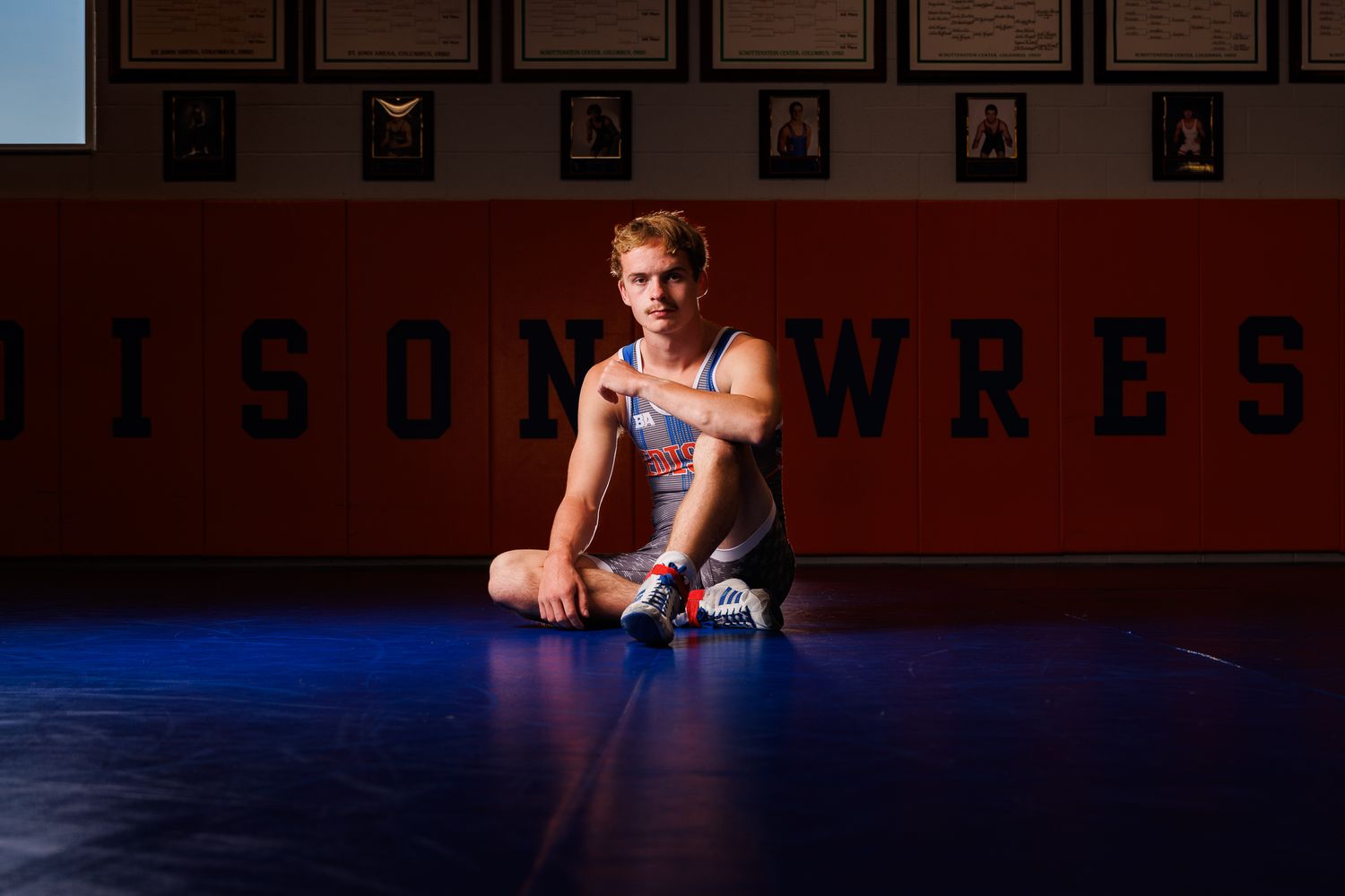 An high school wrestler poses on a wrestling mat wearing a white wrestling singlet under dramatic lighting.