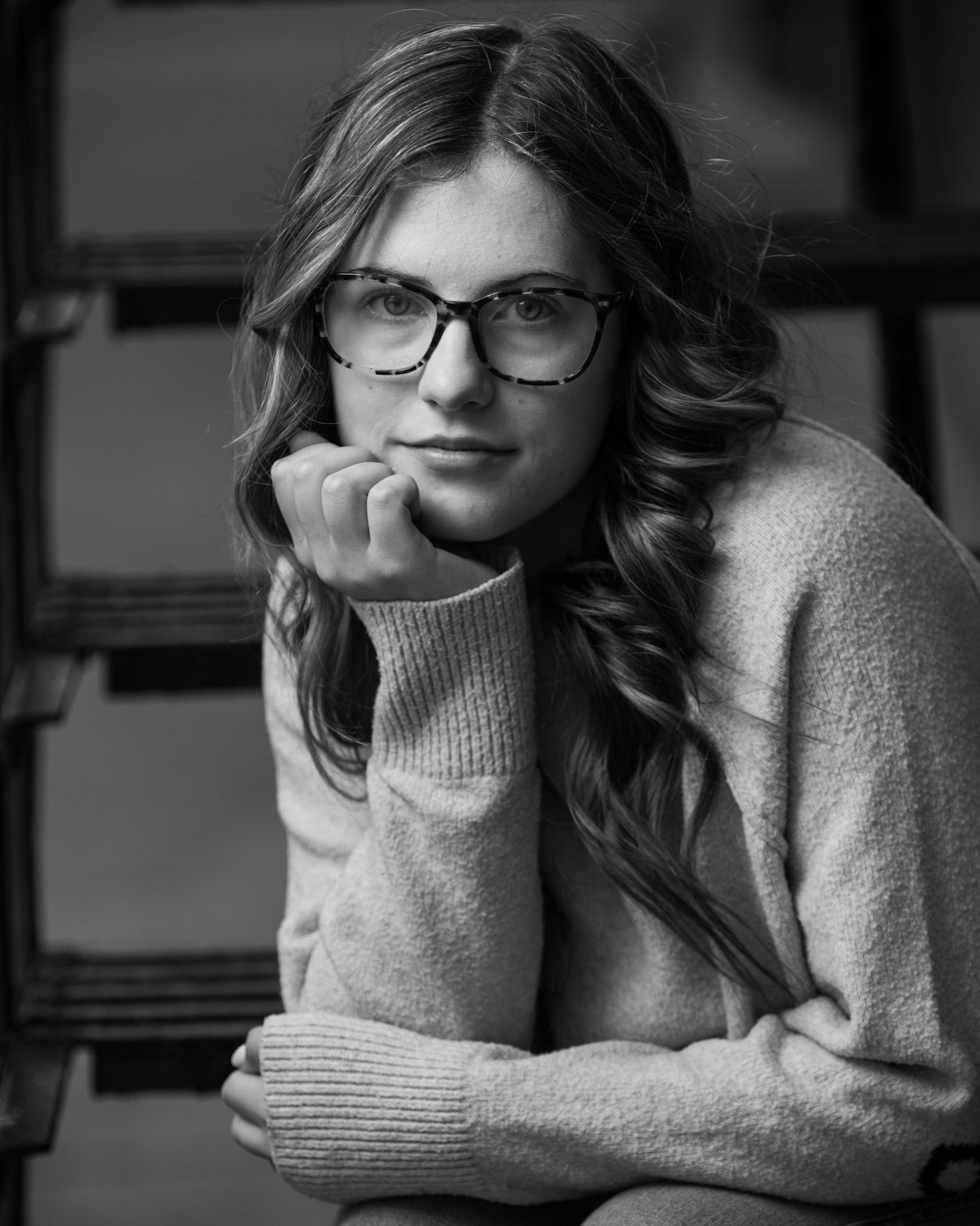 Black and white portrait of someone wearing glasses and a cozy sweater leaning thoughtfully on their hand.