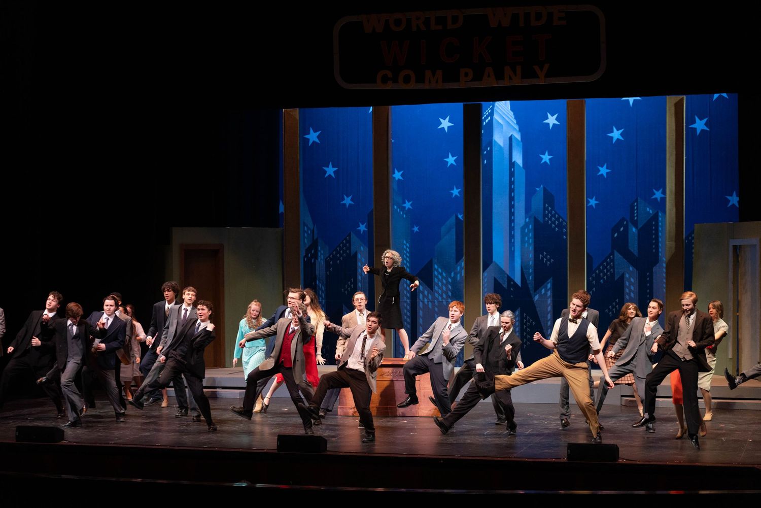 Large ensemble cast performs a lively musical theater number on stage with a city skyline backdrop and star decorations.