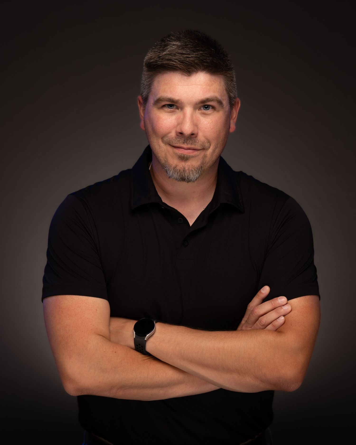 Professional headshot of a confident man in a black polo shirt with arms crossed against a dark background.