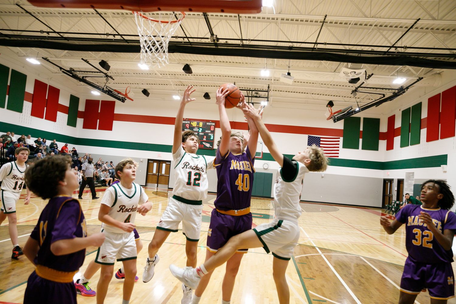 High school basketball game with players in purple and white uniforms competing for a rebound in a green and red decorated gym.