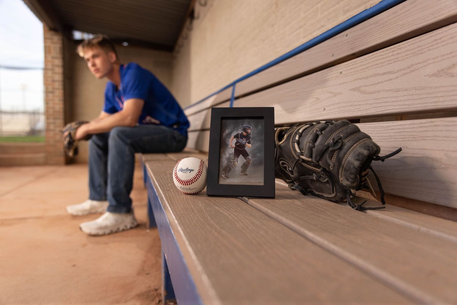 Teen baseball player sits in dugout beside a framed photo, baseball, and worn glove as a memorial tribute.