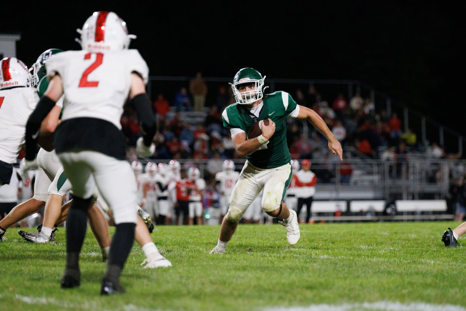 High school football player in green uniform runs with the ball during a night game against red and white opponents.