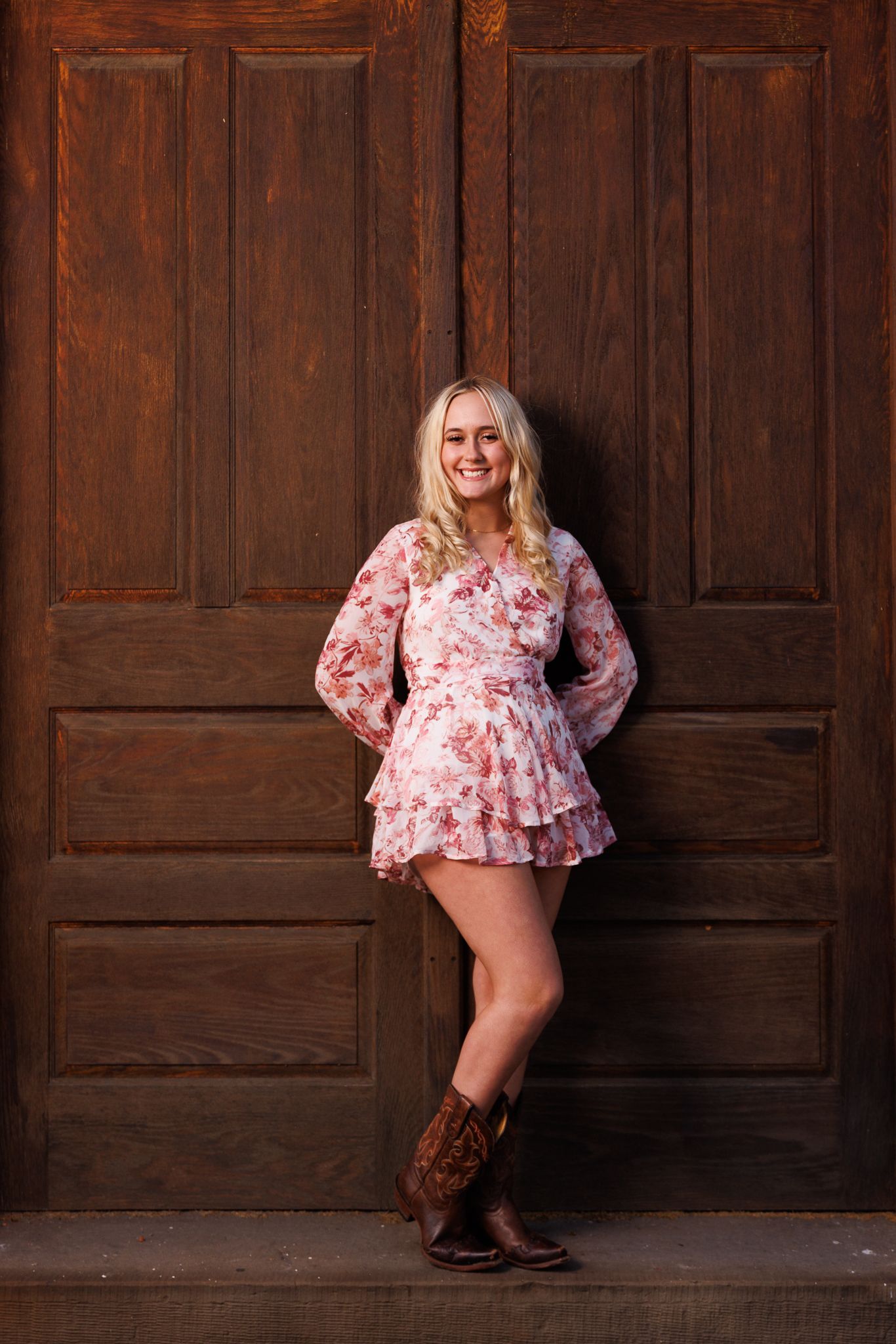 Young woman in floral pink mini dress and brown cowboy boots poses against rustic wooden door.