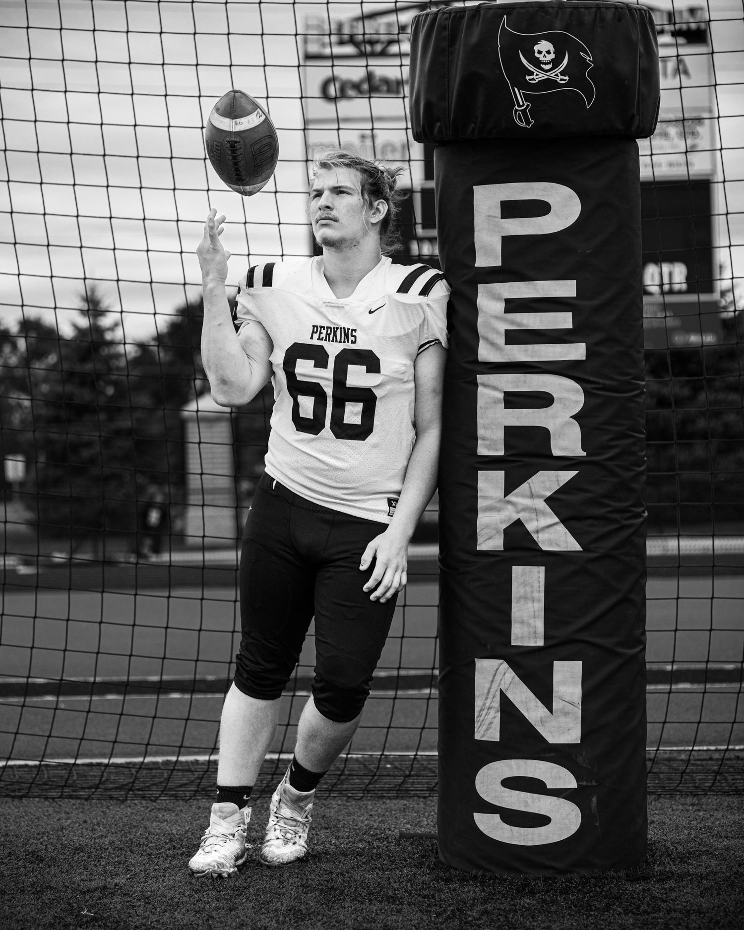 A black and white image of a high school football player tossing a football while leaning against a padded goal post.