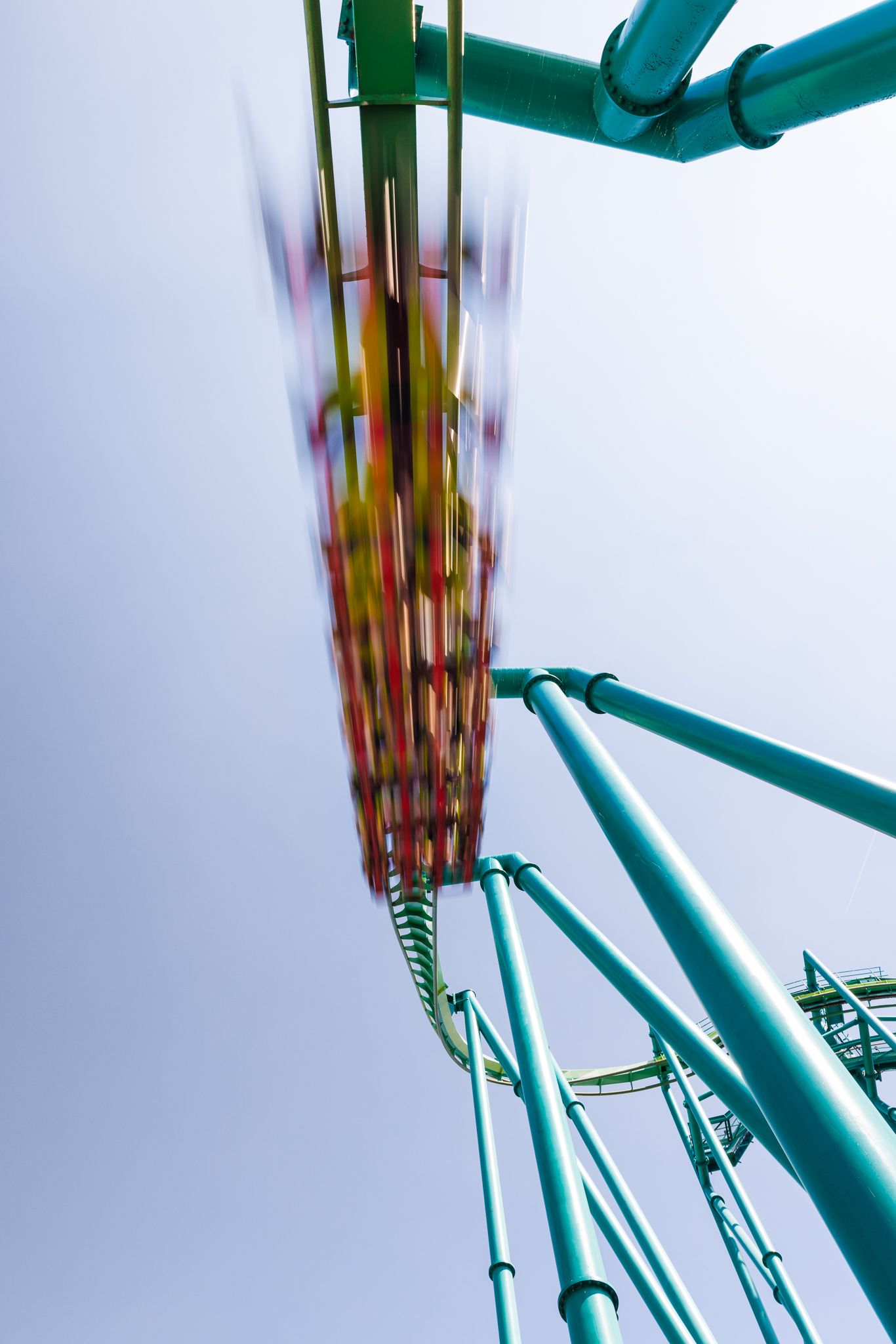 Roller coaster cars speeding down a steep drop on teal track, captured with motion blur against a pale sky.