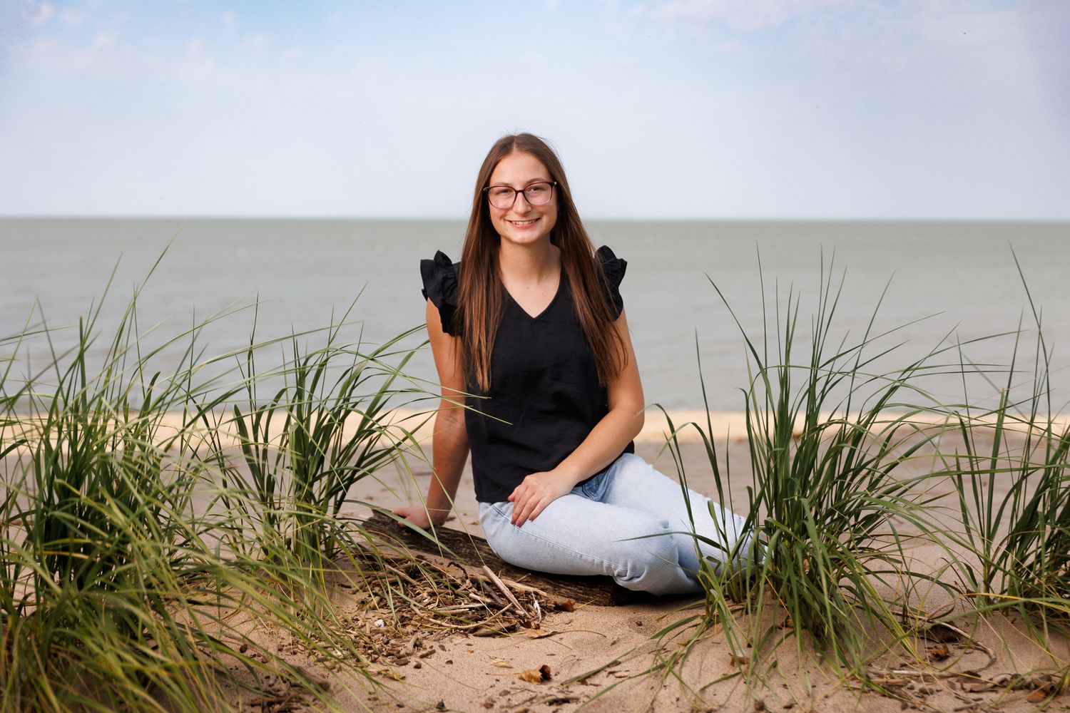 A high school senior in casual attire sits and poses by beach grass by Lake Erie at sunset.