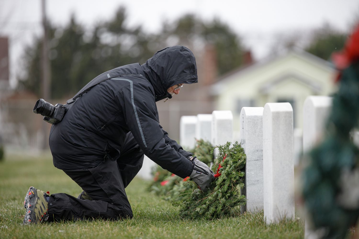 Person in black winter gear kneeling at military gravestone placing holiday wreath in cemetery.