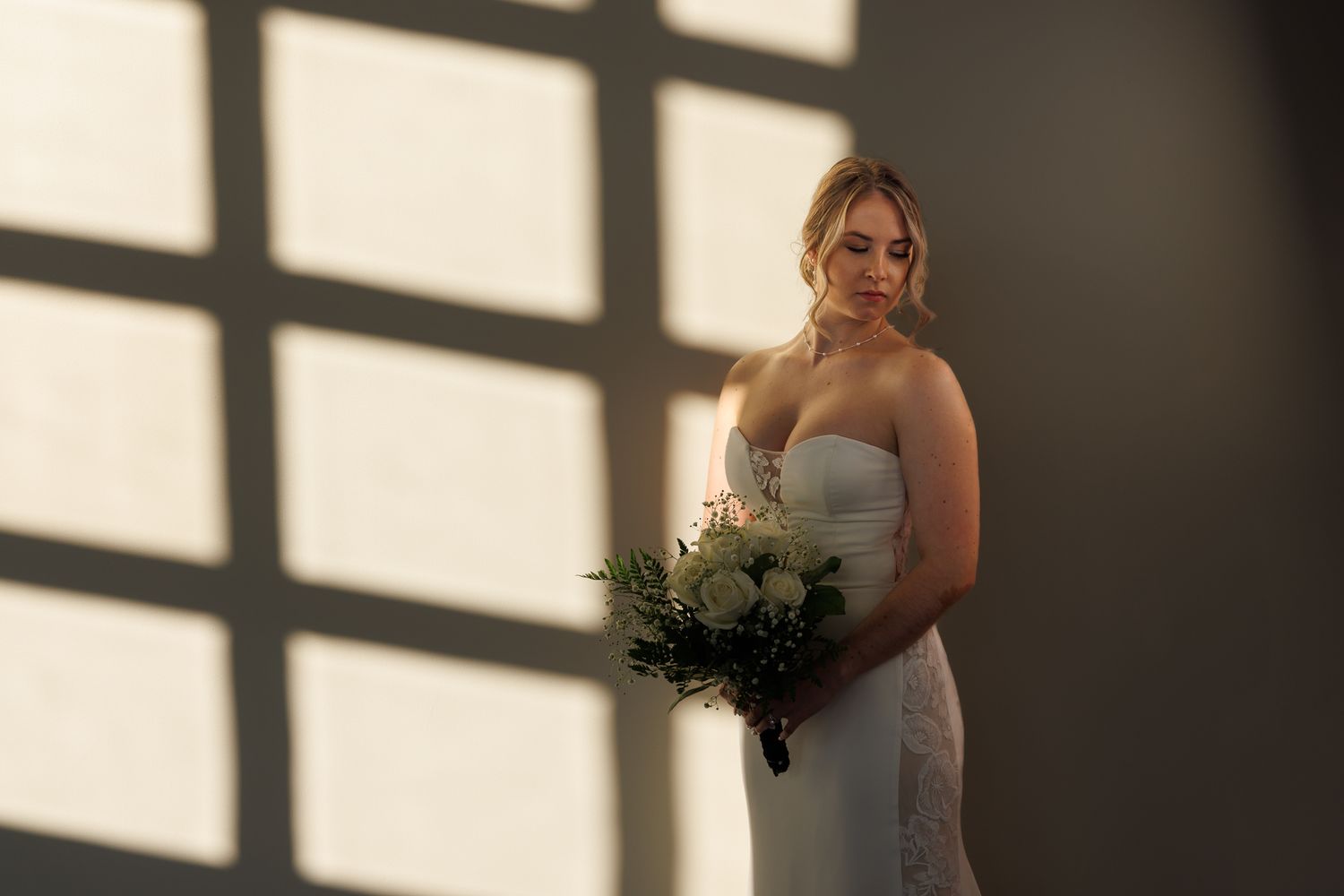 Bride in strapless white gown holding bouquet of white roses, bathed in soft window light against a neutral wall.