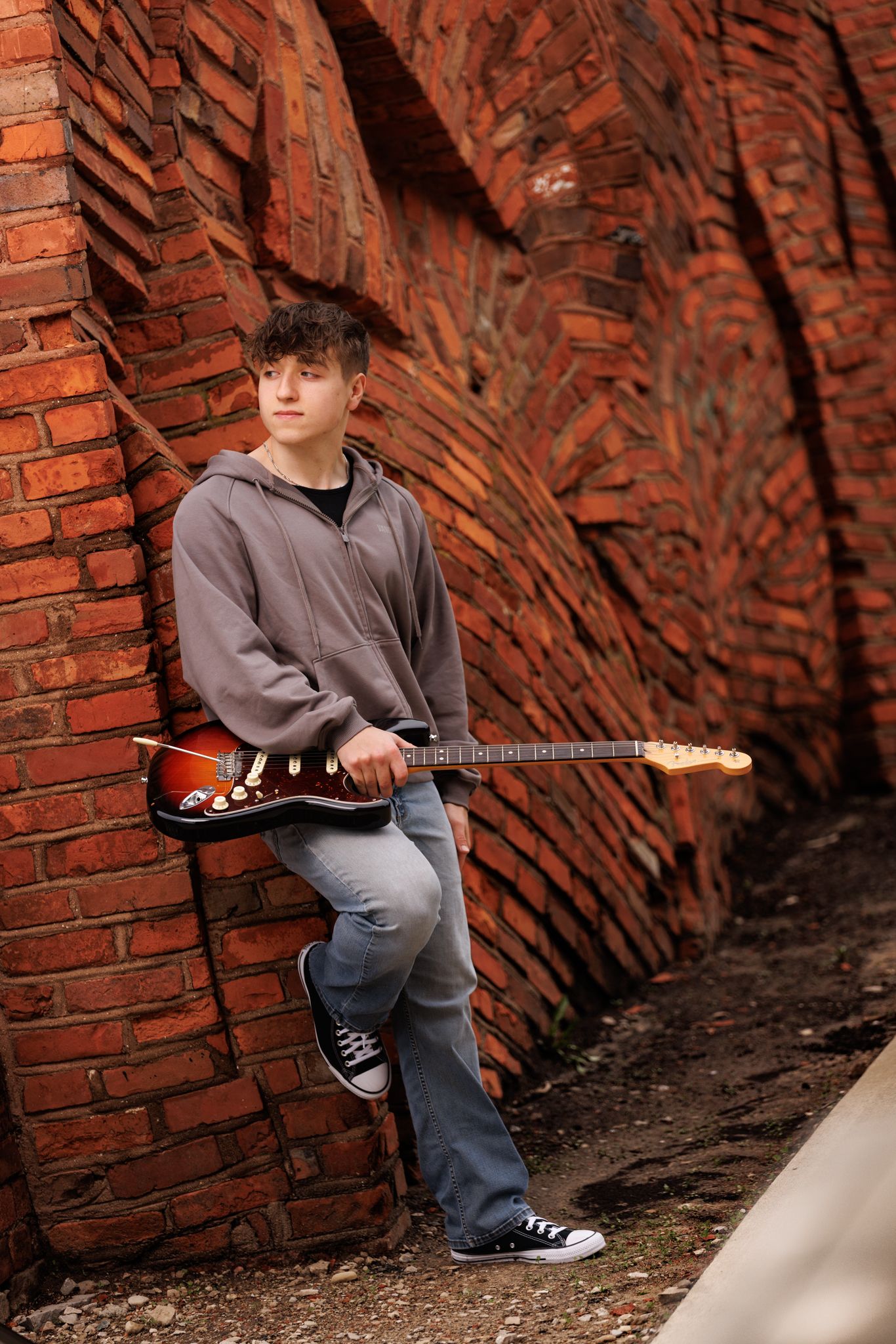 A musician poses with an electric guitar against a textured red brick wall in casual attire in historic downtown Sandusky, Ohio.