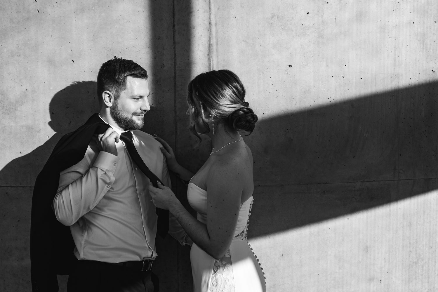 Black and white photo of a bride and groom adjusting his suit jacket against a wall with dramatic shadows.