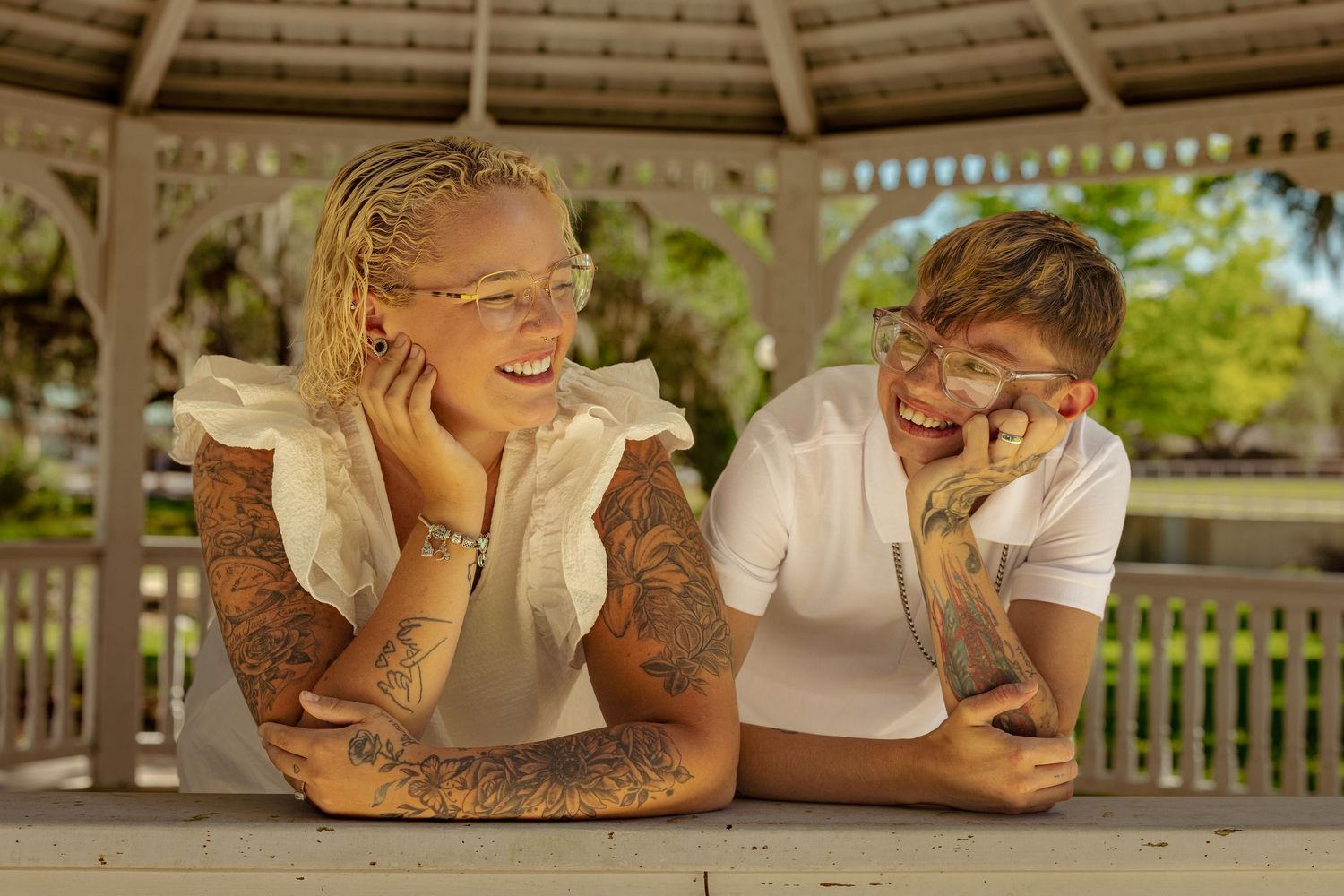 Two tattooed women laughing together under a gazebo in a sunny park setting.