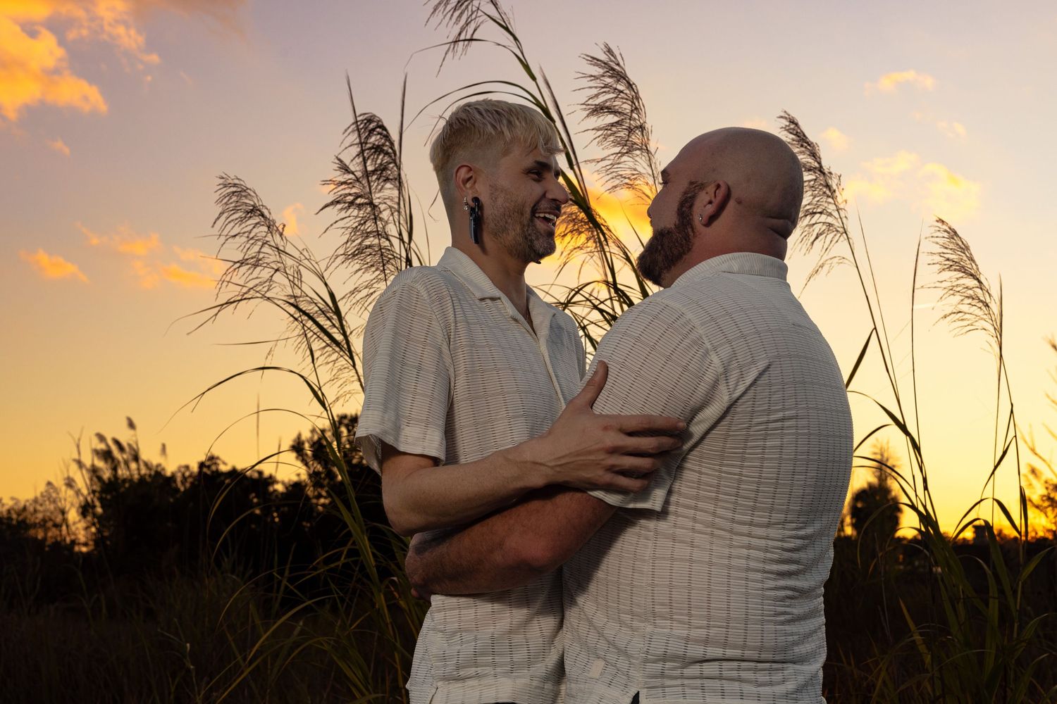 Two men embrace romantically at sunset in a field of tall grasses, sharing an intimate moment outdoors.