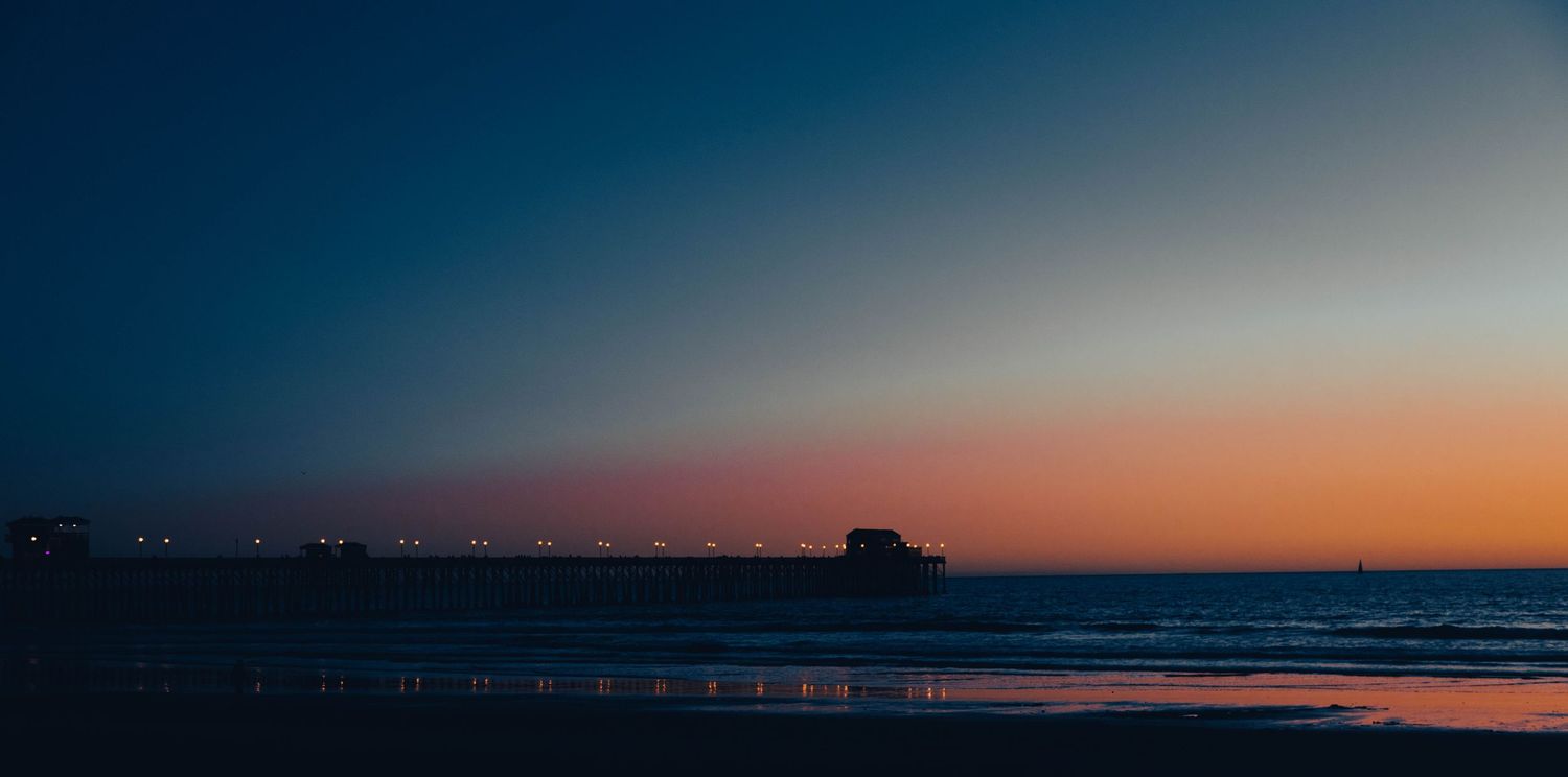 Stunning twilight beach scene with glowing pier lights reflecting on wet sand under a vibrant orange and blue sunset sky.