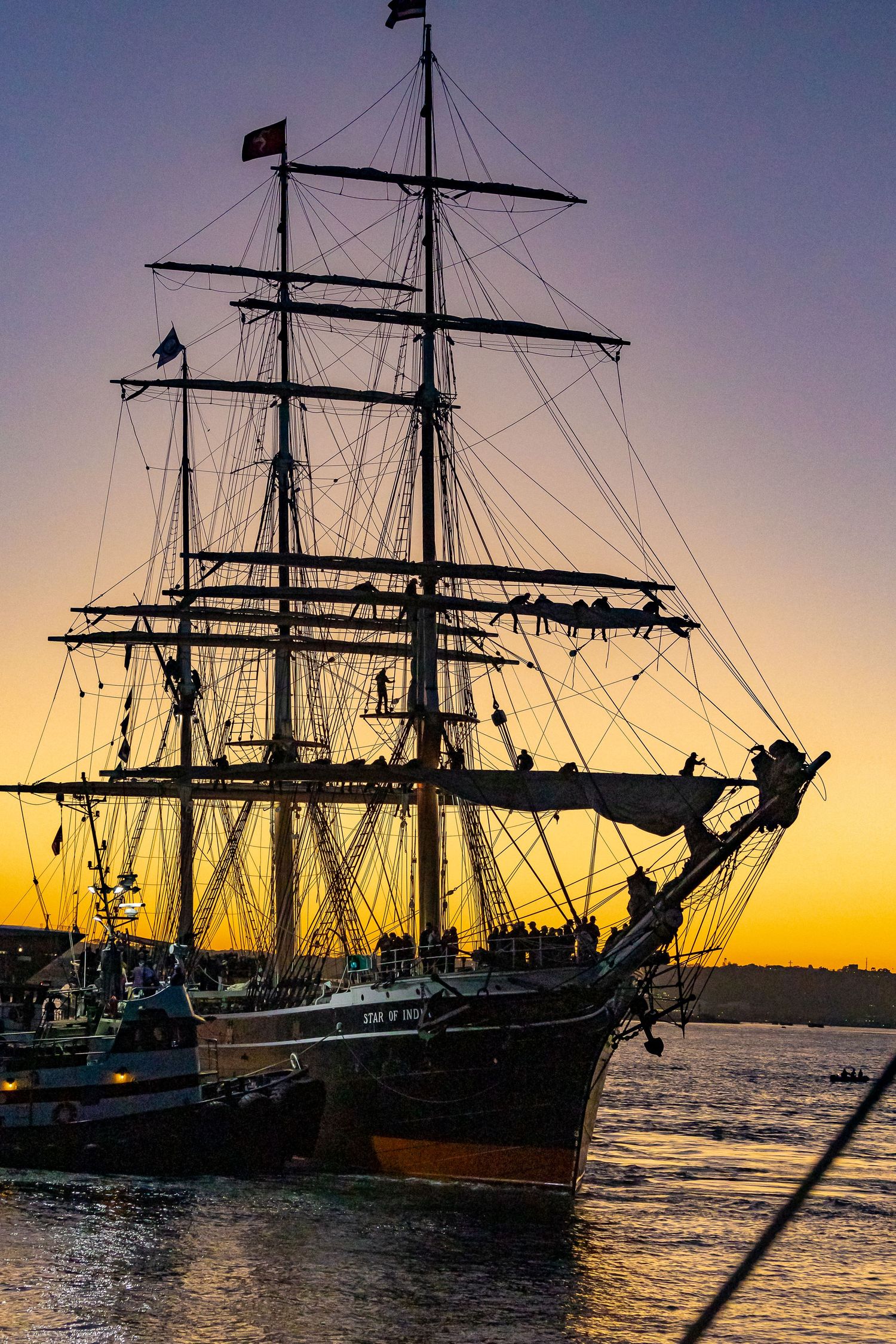 Tall ship silhouetted against a golden sunset sky, moored in calm harbor waters with intricate rigging and masts visible.
