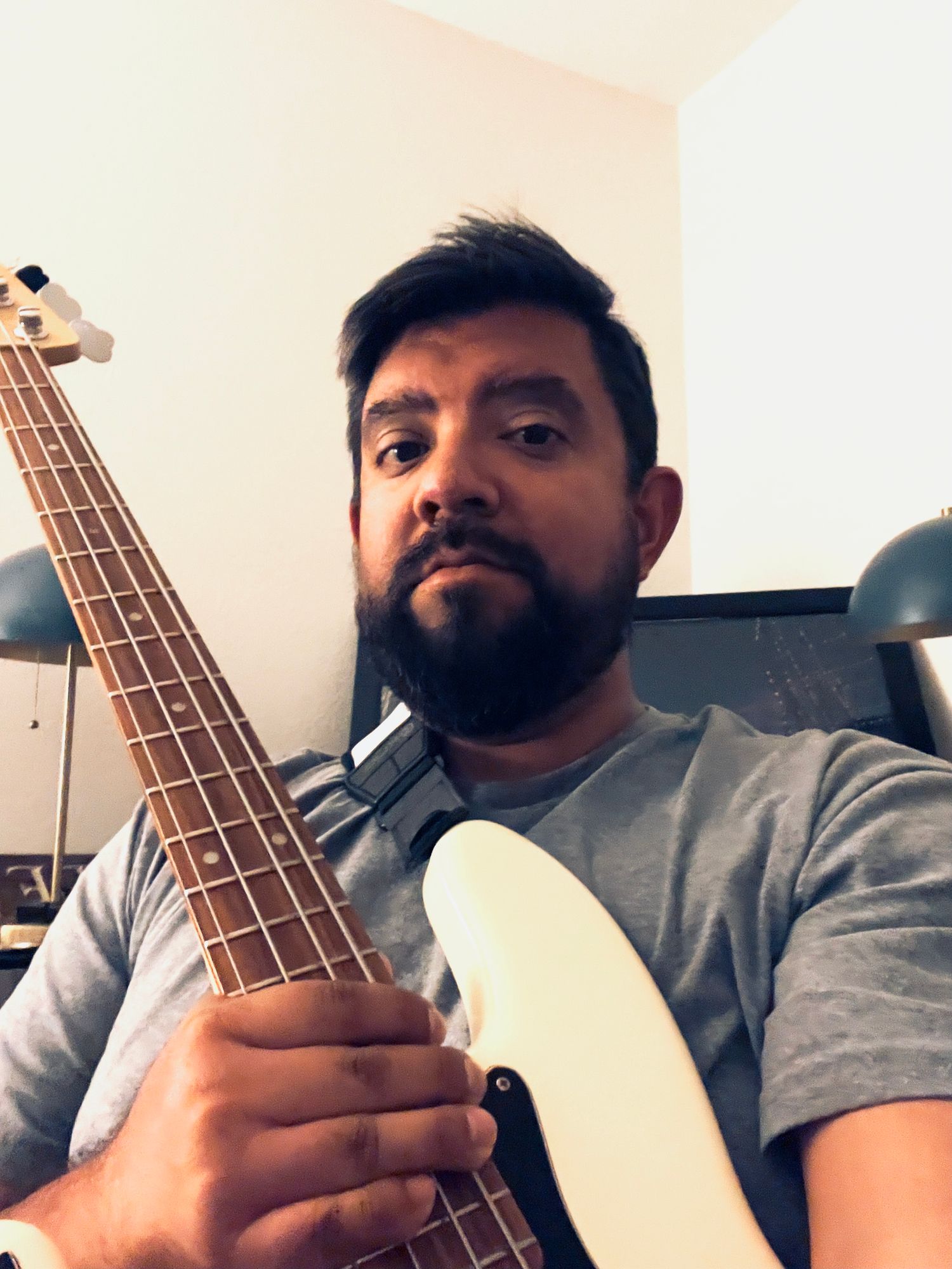 Man with beard holding a white bass guitar indoors, wearing a gray t-shirt, with studio lighting in background.