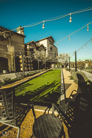 Outdoor winery patio with string lights, bocce ball court, wooden chairs, and wine barrels under a clear blue sky.