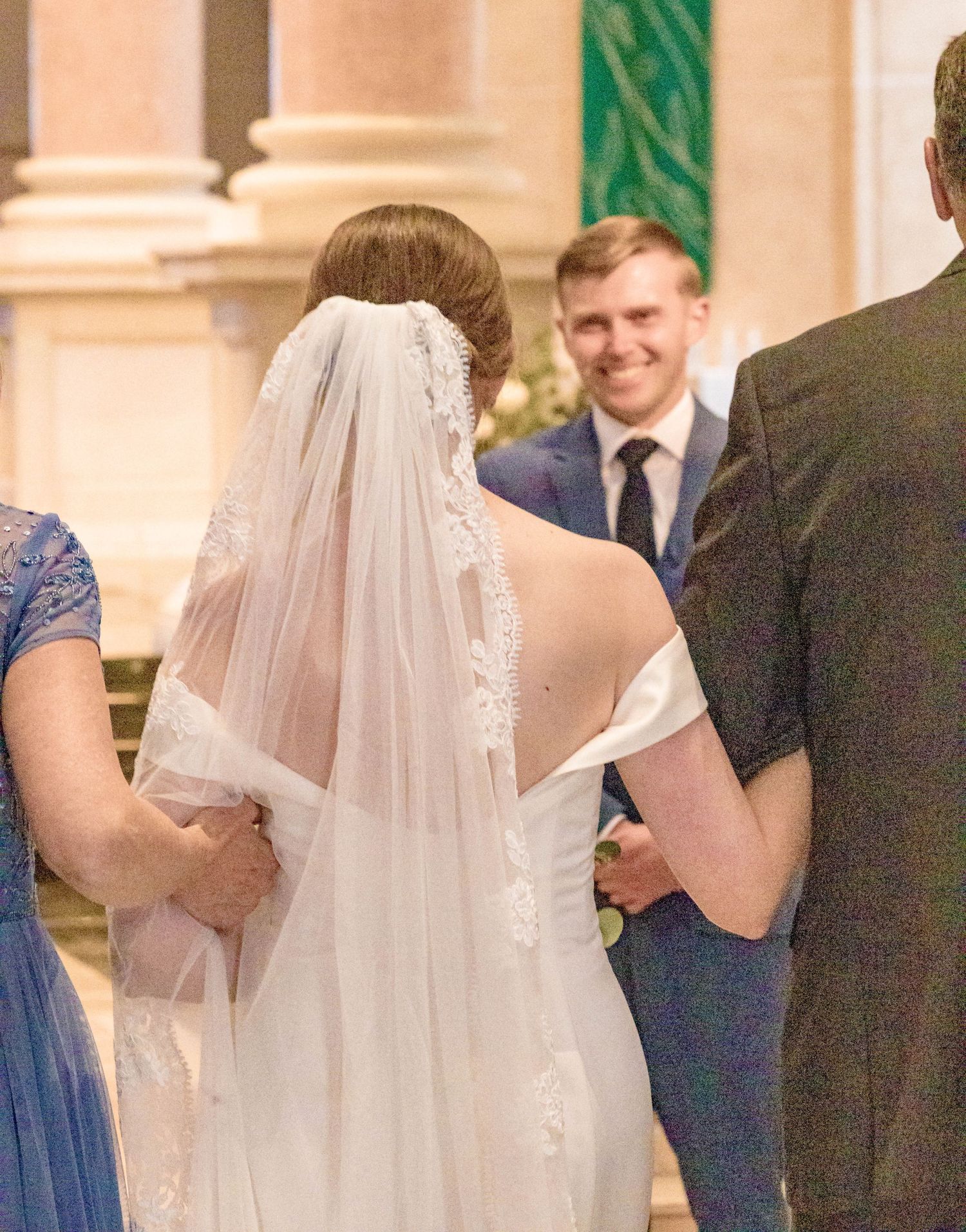 Bride in white off-shoulder gown and lace veil at church wedding ceremony with smiling groom in navy suit.