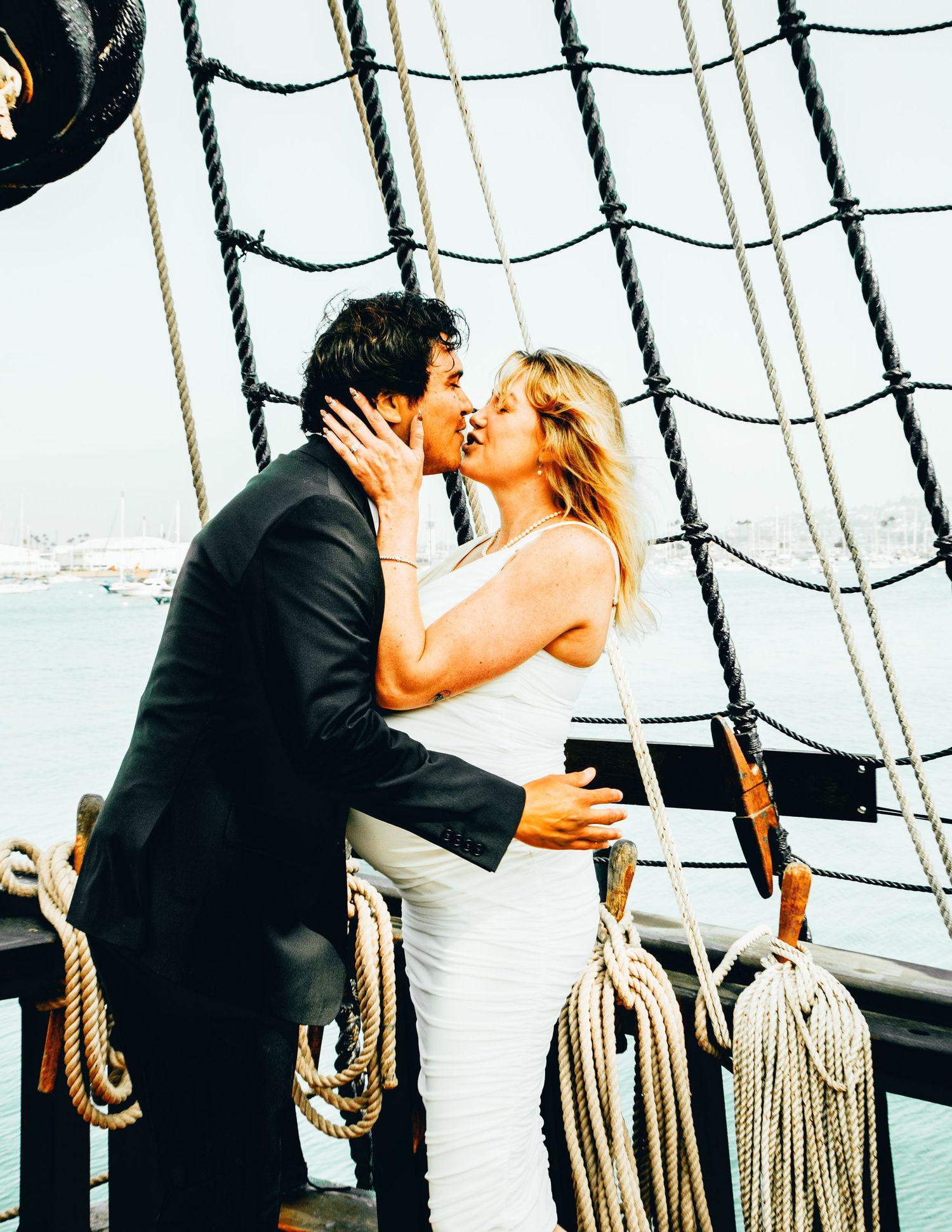 Couple sharing a romantic kiss on a tall ship deck, woman in white dress, man in black suit, with rigging and harbor backdrop.