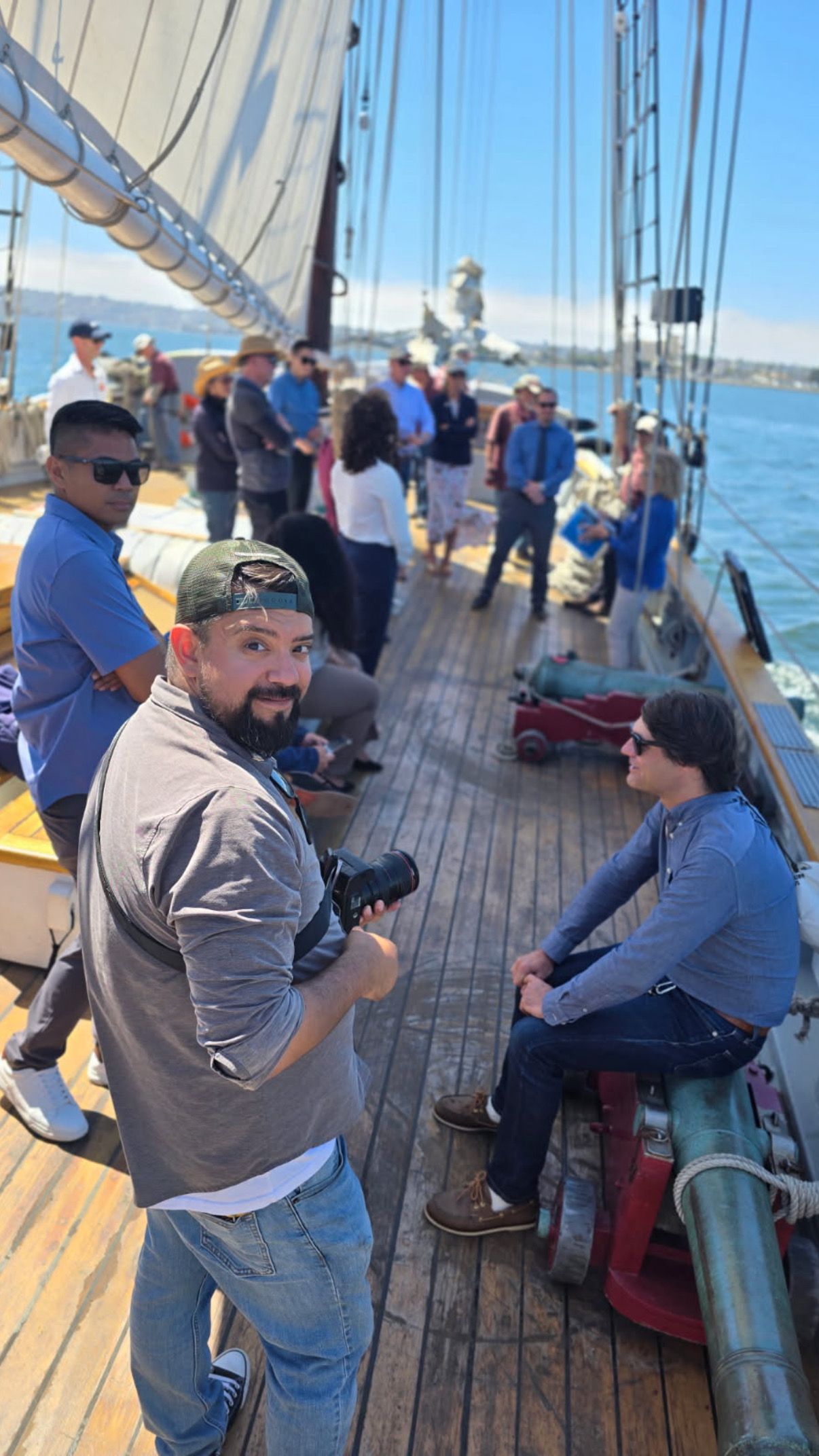 Photographer holding camera on a wooden tall ship deck with passengers enjoying a sunny sailing excursion on the bay.
