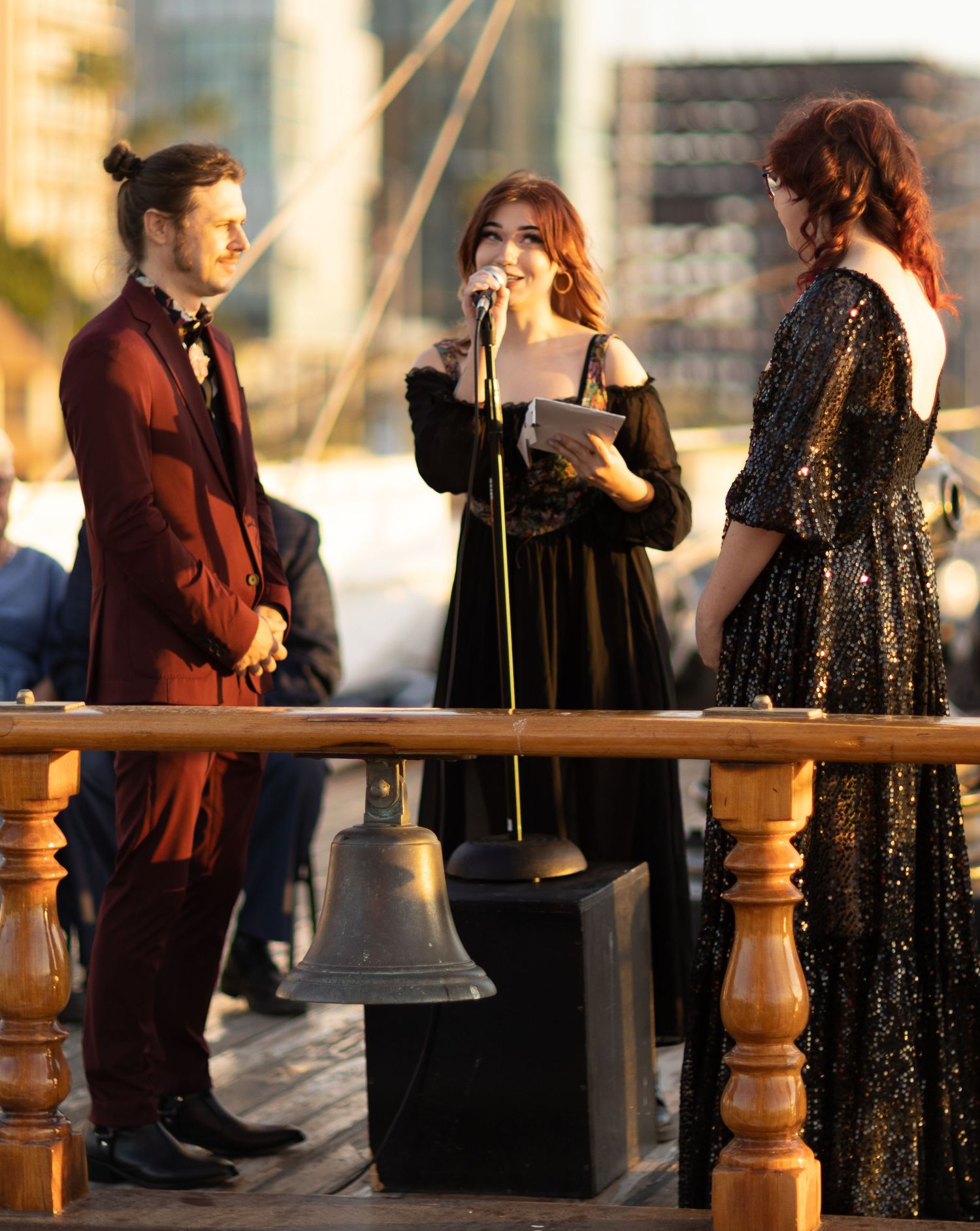 Elegant outdoor wedding ceremony on a dock with three people in formal black and burgundy attire near a ship's bell.