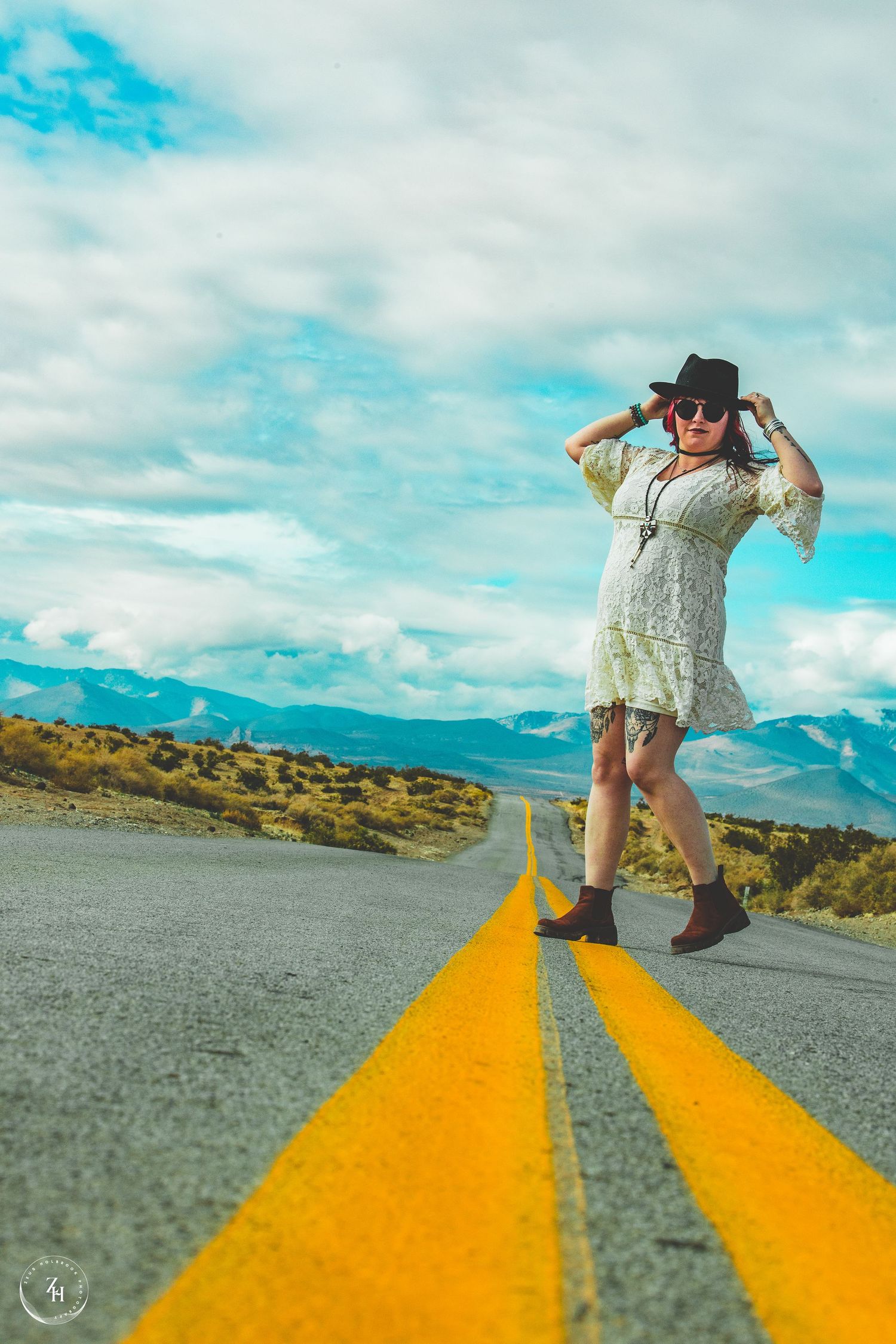 Woman in white dress and black hat poses on a open road with yellow lines, mountains and cloudy sky in background.