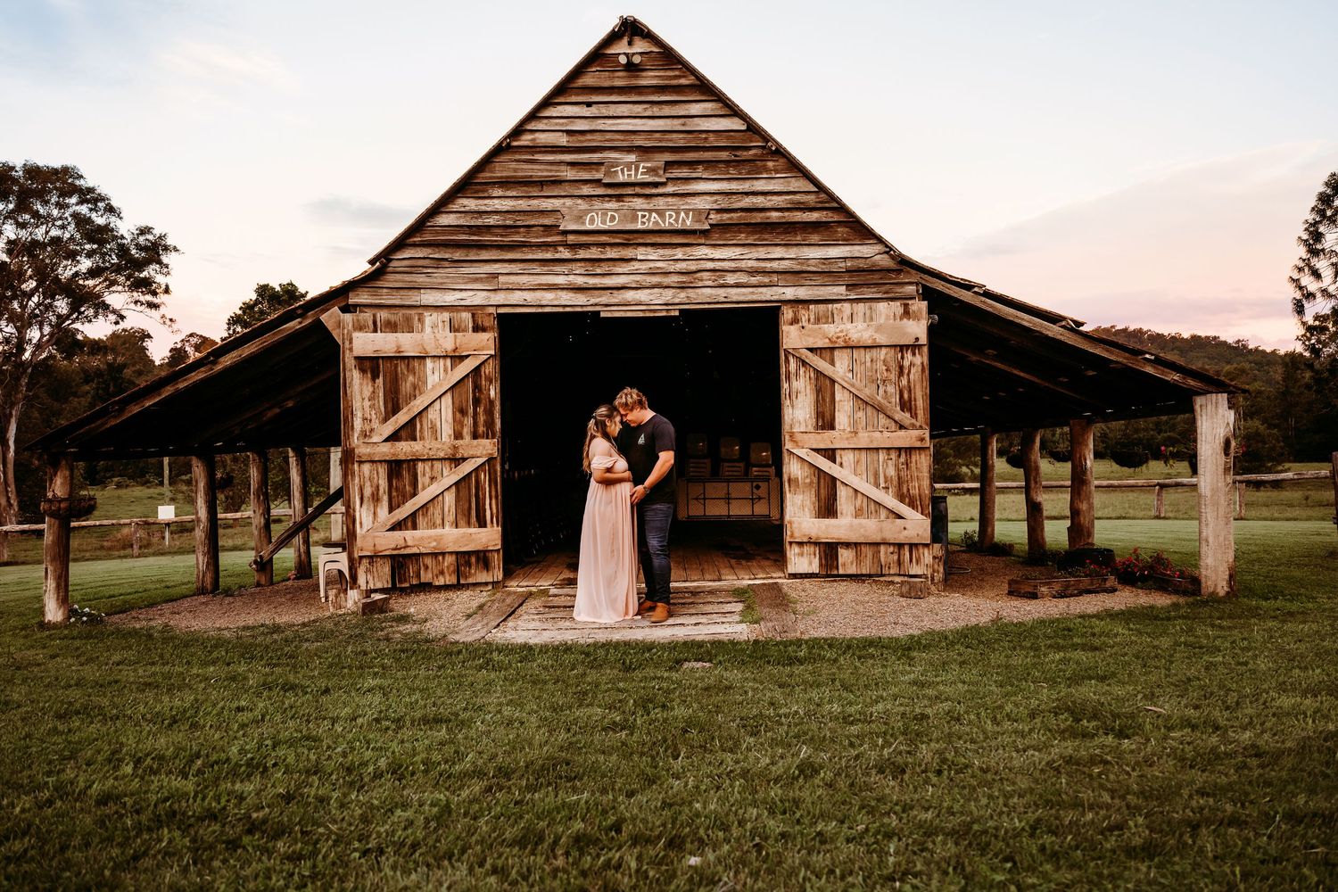 A rustic wooden barn with open doors at sunset creates a romantic silhouette against the pastoral landscape.