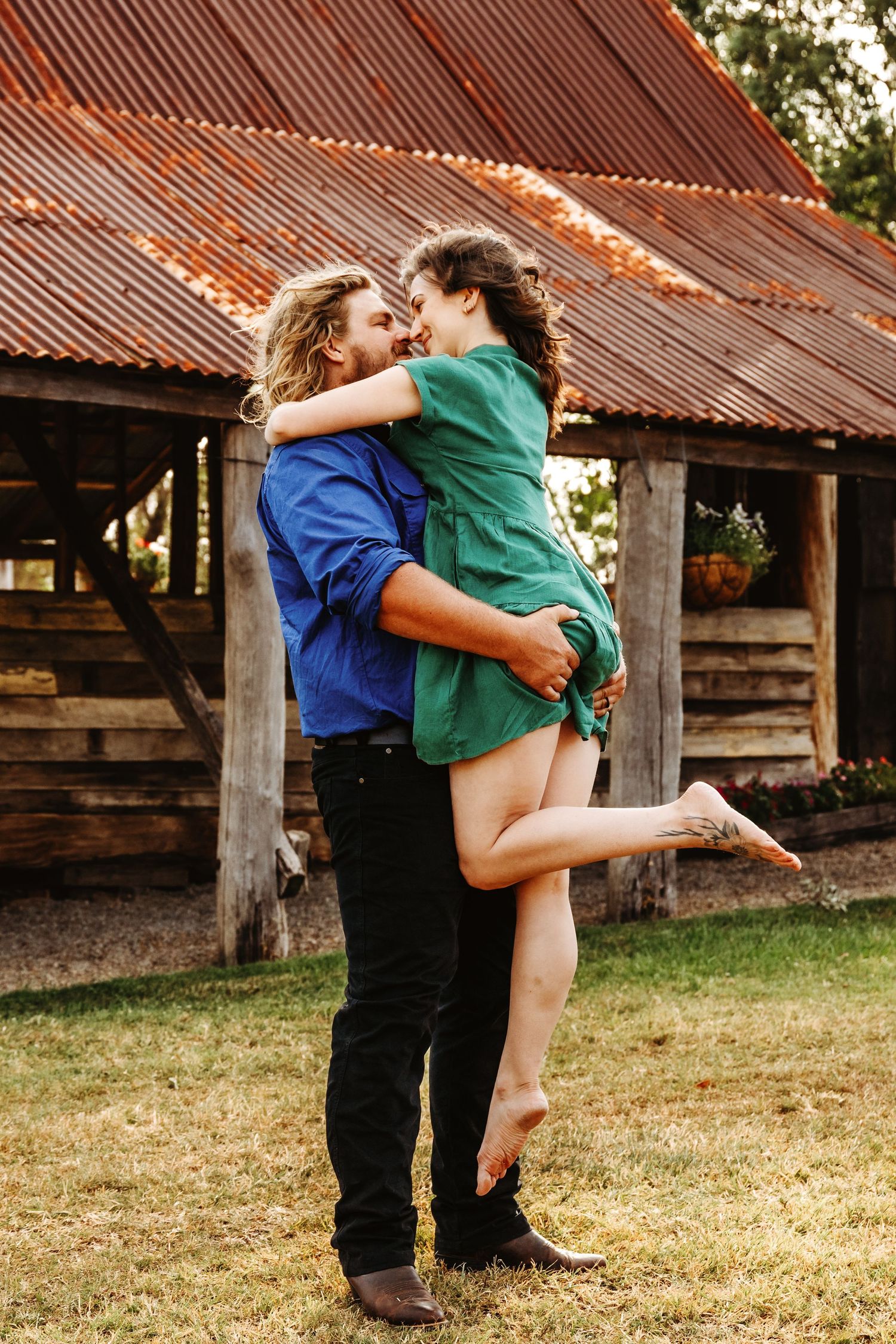 A romantic embrace between a couple in casual clothing standing in front of a rustic barn with a metal roof.