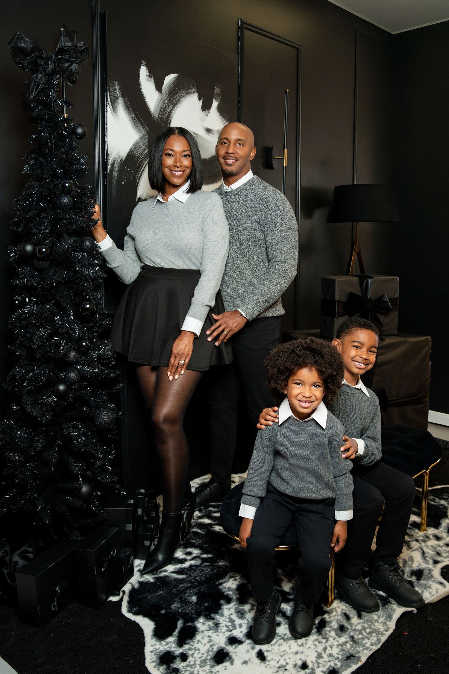 Family of four posing elegantly in coordinated gray outfits beside a black Christmas tree in a dark, modern room.