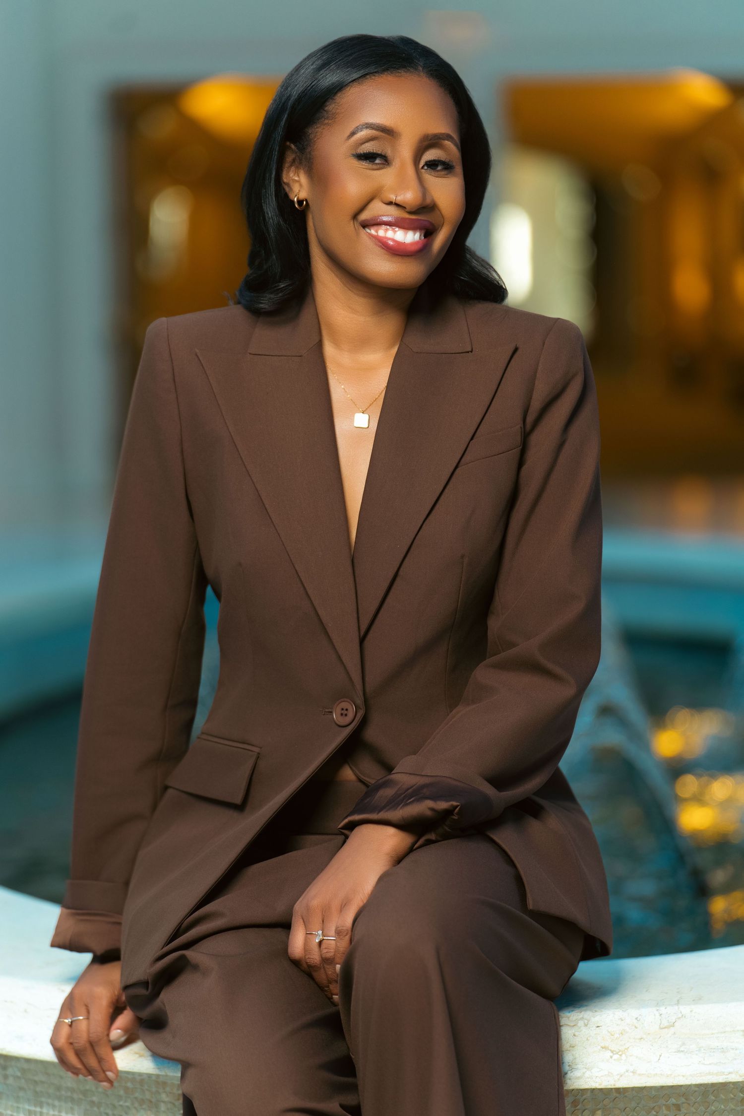 A smiling woman in a brown blazer suit sits confidently by a fountain in an elegant indoor setting.