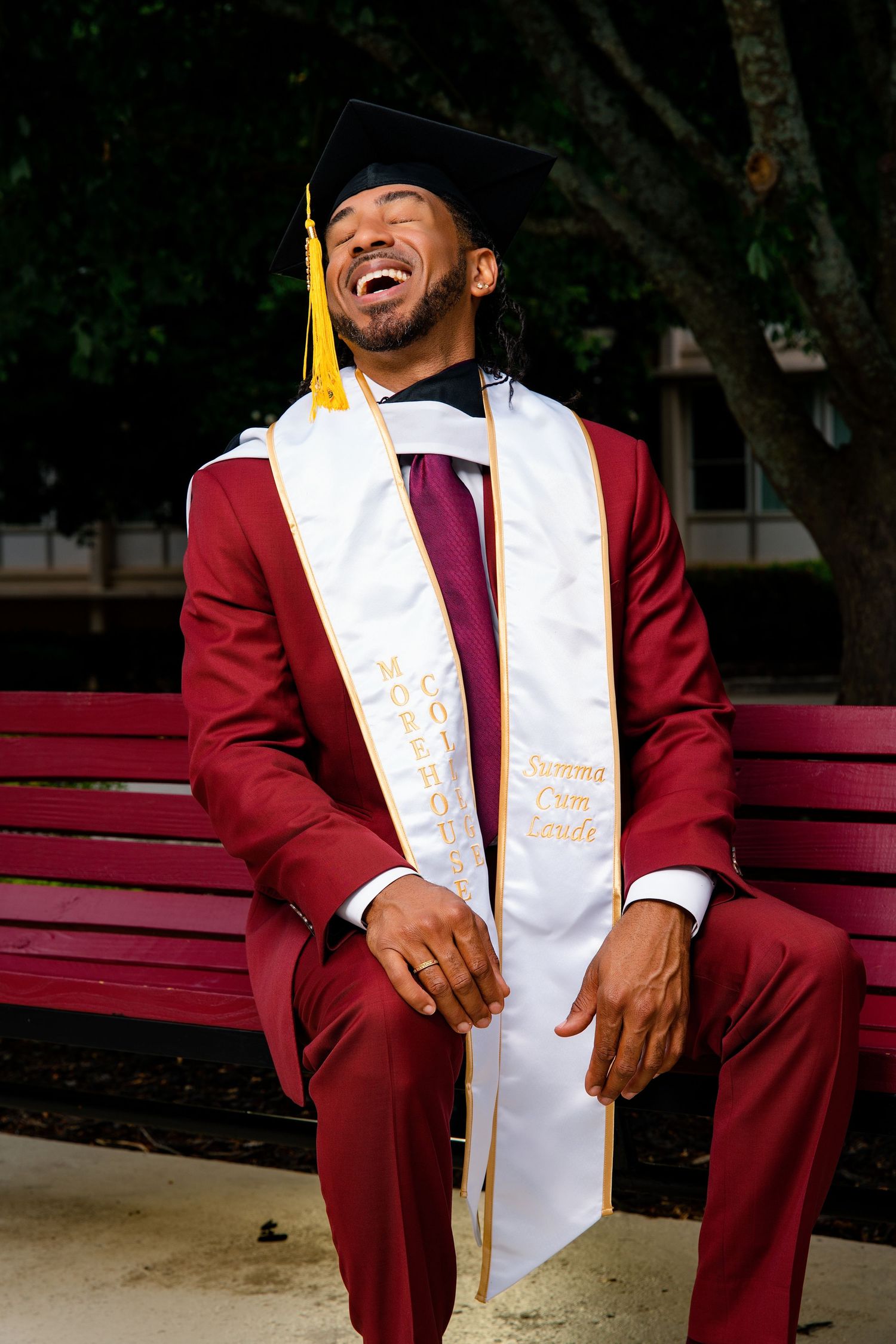 Joyful Black man in maroon graduation suit, cap, and Summa Cum Laude stole sits laughing on a bench.