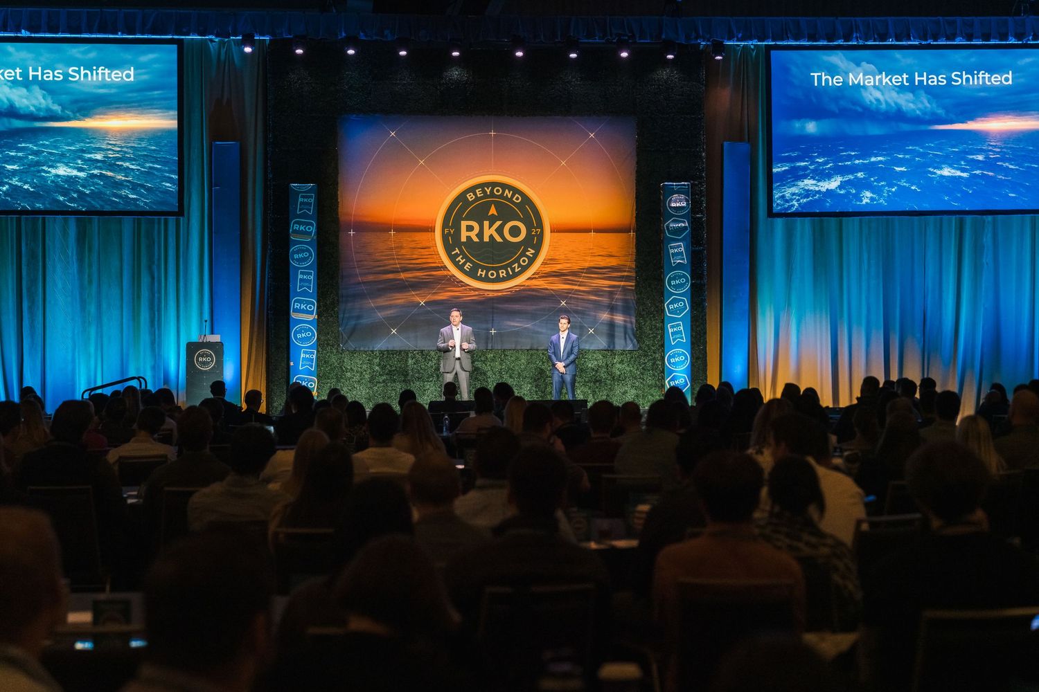 Large corporate conference with audience seated before a stage displaying RKO logo and 'The Market Has Shifted' presentation.