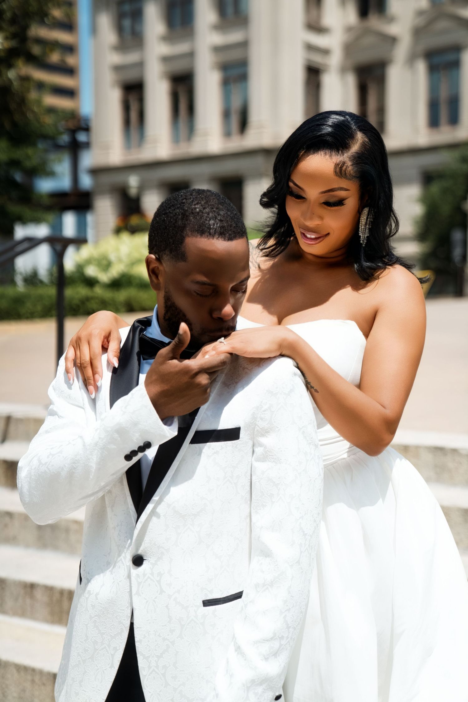 A groom in a white tuxedo kisses his bride's hand as she smiles, both dressed in elegant wedding attire outdoors.