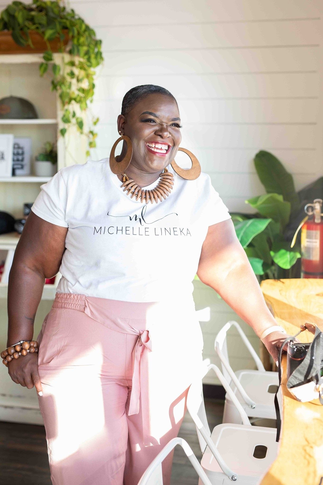 A joyful person wearing a white t-shirt and pink pants stands in a bright kitchen with plants and decor.