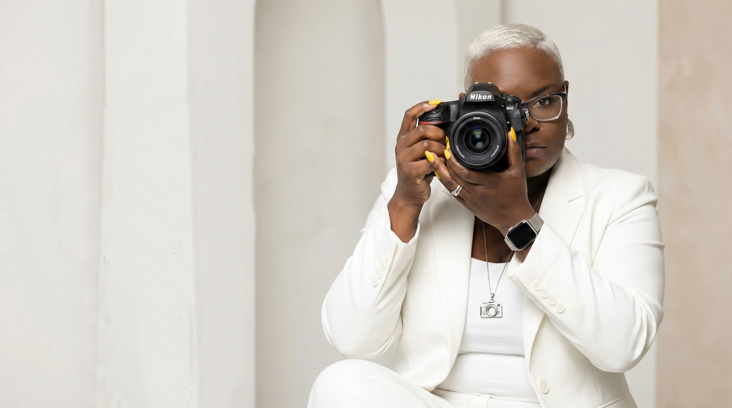 A Black woman in a white blazer holds a Nikon DSLR camera up to her face, photographing indoors.