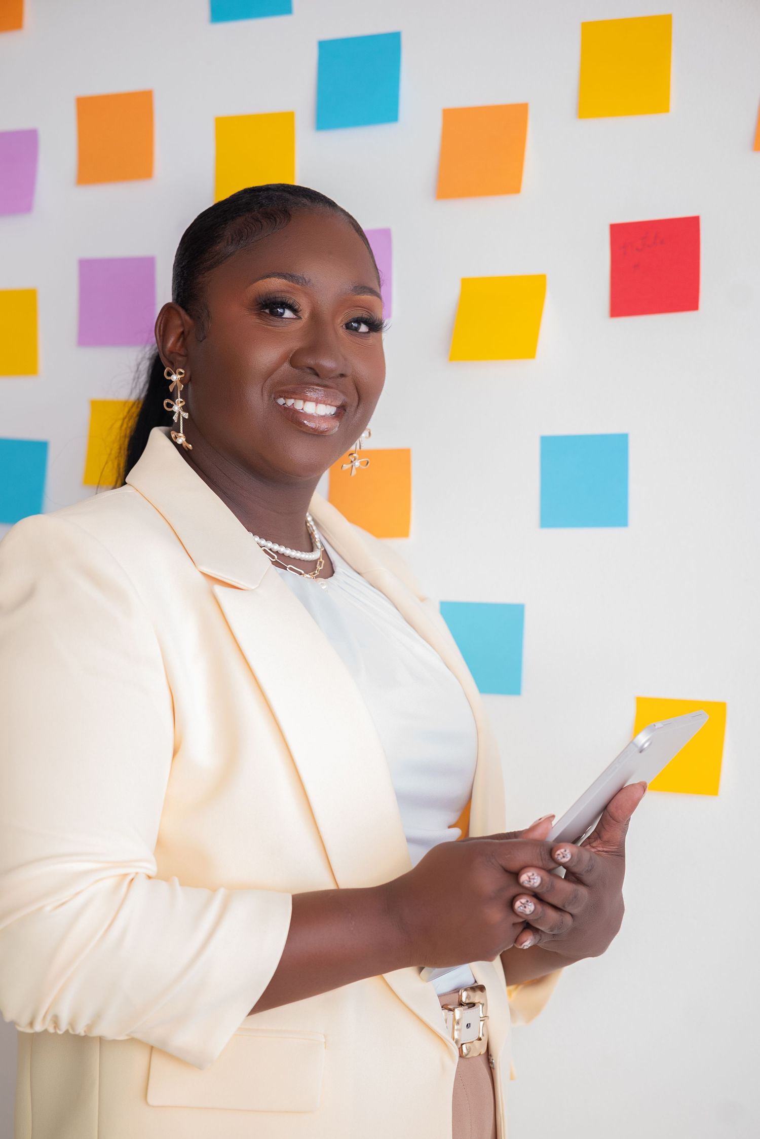 Smiling businesswoman in cream blazer holding tablet, standing in front of colorful sticky notes on white wall.