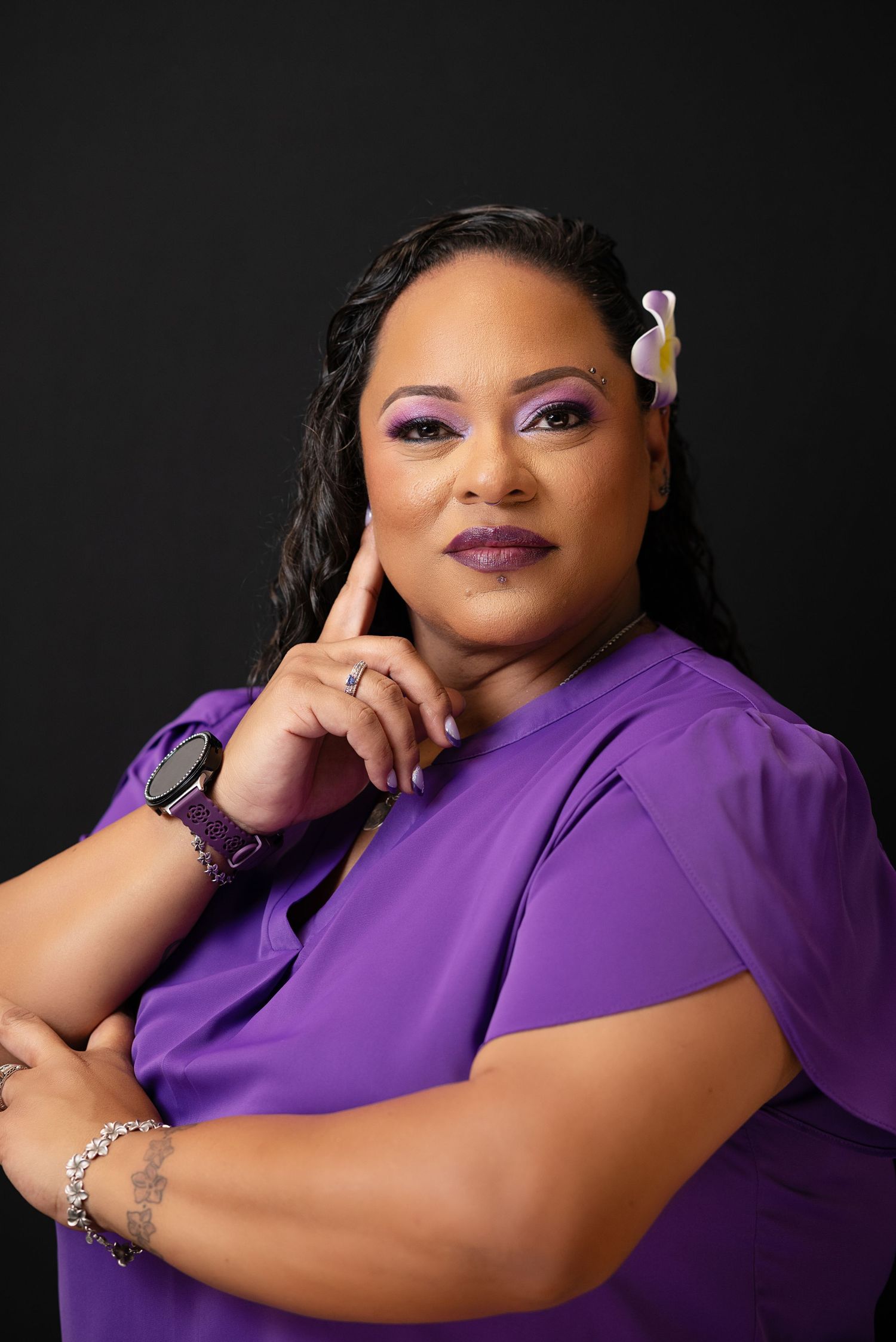 A confident woman in a purple outfit with matching makeup and a floral hair accessory poses thoughtfully against a dark background.