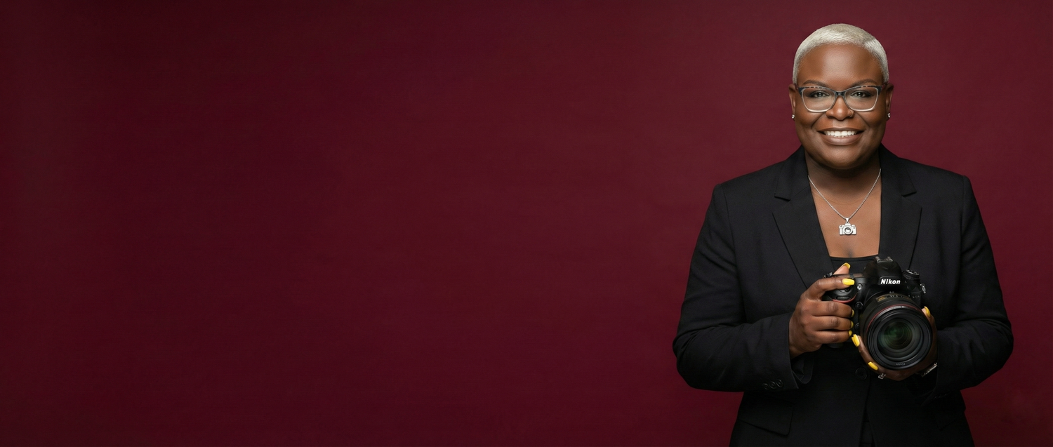 A person in a black blazer smiles while holding a microphone against a deep red background.