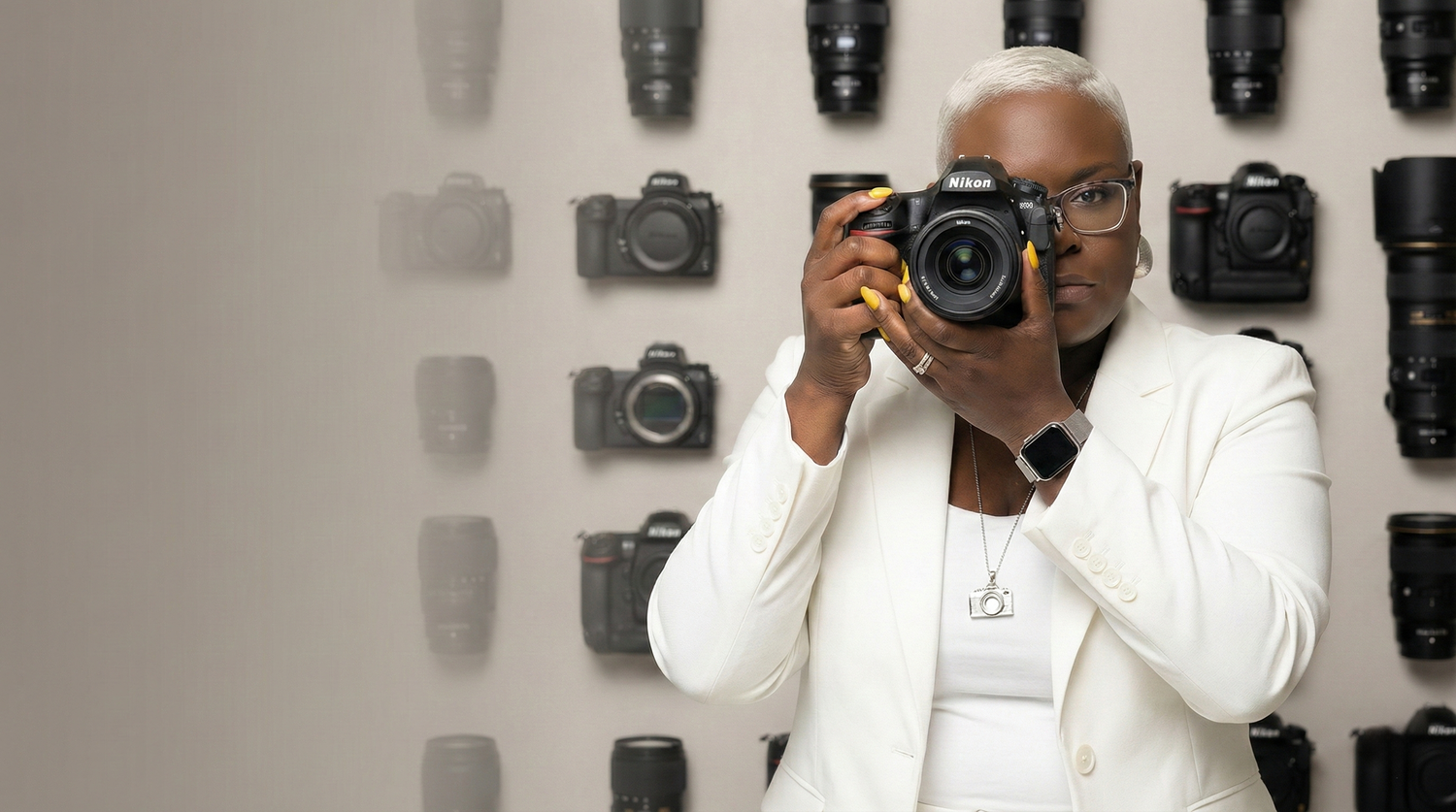 A photographer holds up a black DSLR camera while standing against a white background.