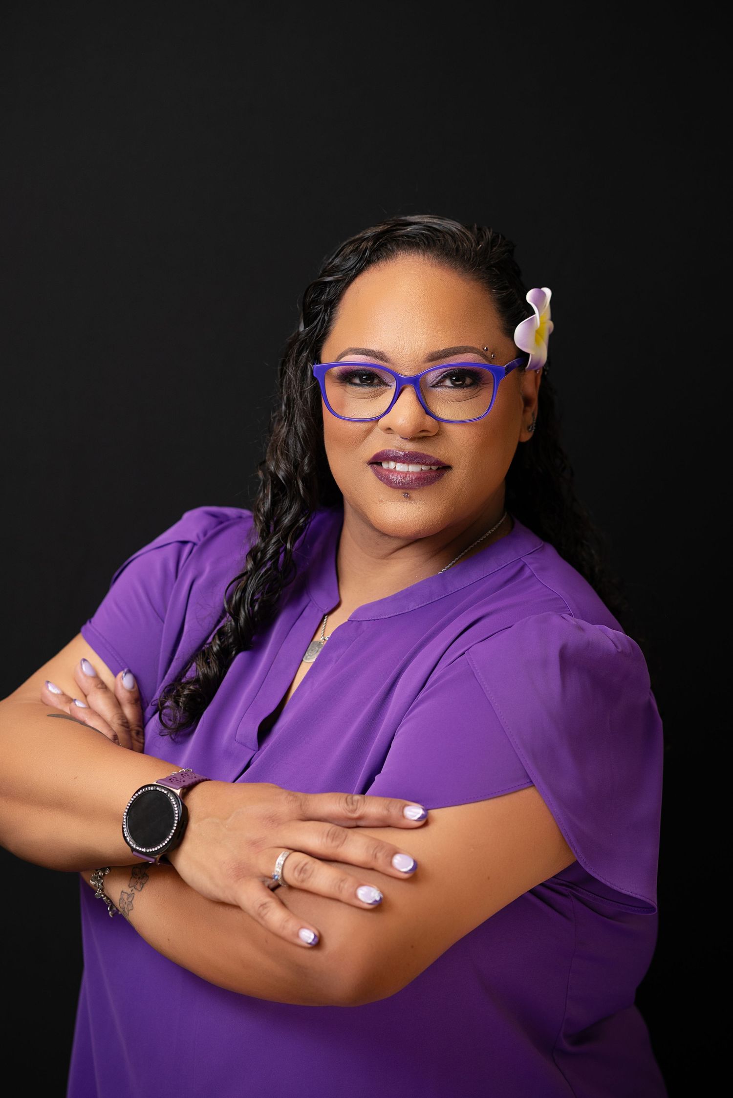 Woman in purple top and glasses with flower hair accessory poses confidently with arms crossed against dark background.