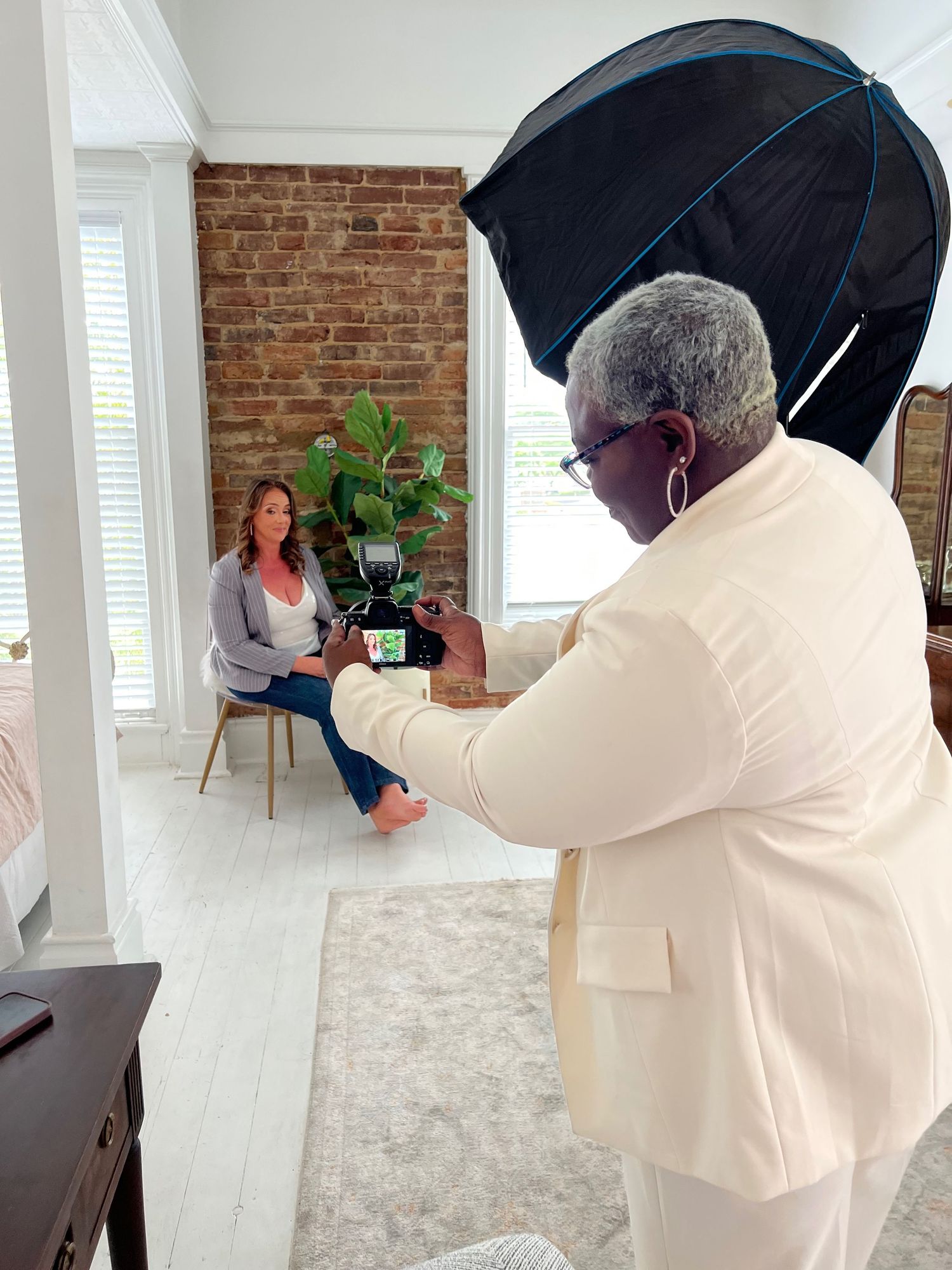 Photographer with umbrella light photographs a woman seated on a chair in a bright, brick-walled studio.