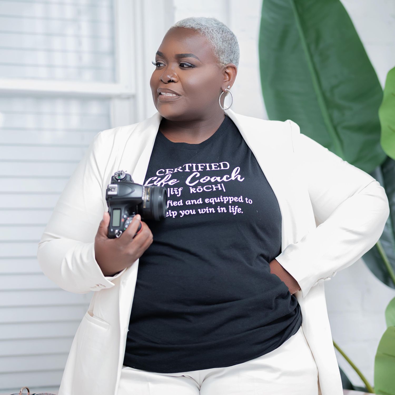 Woman in white blazer and black certified life coach t-shirt holding a camera, posing confidently indoors.