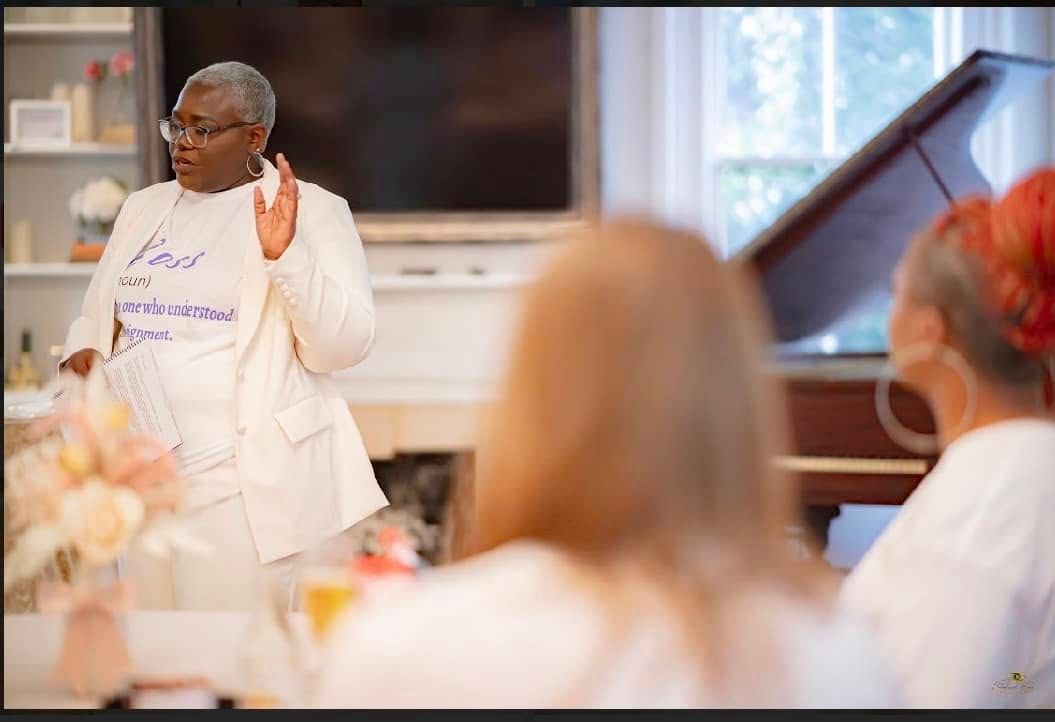 A person in a white lab coat gestures while speaking to a seated audience in an elegant room with a piano.