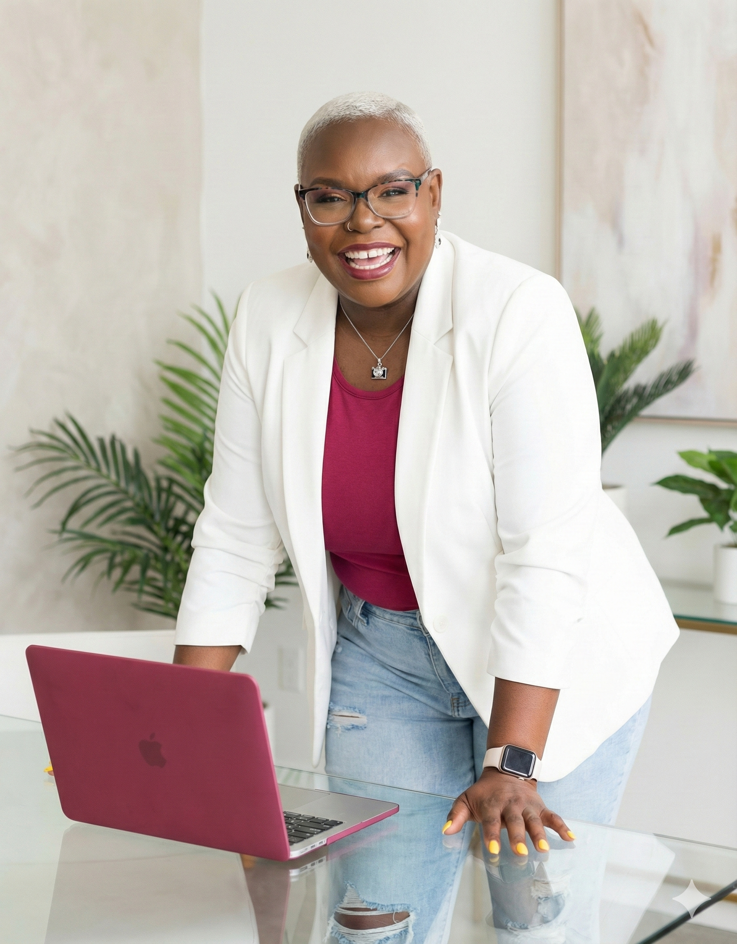 Smiling woman in white blazer and red top leans on glass desk with pink laptop in bright modern office.