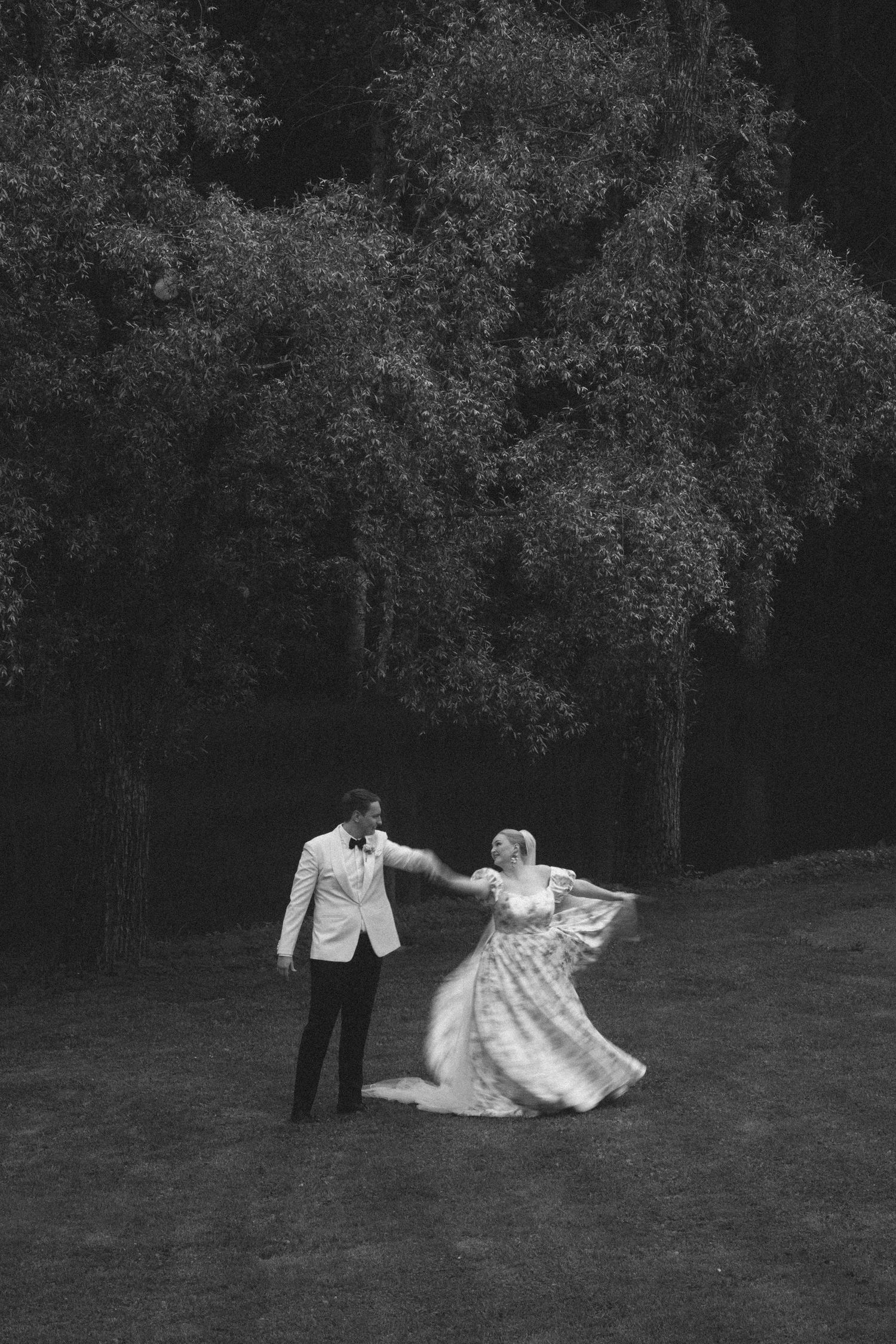 Black and white photo of a bride and groom dancing outdoors on a lawn with lush trees in the background.