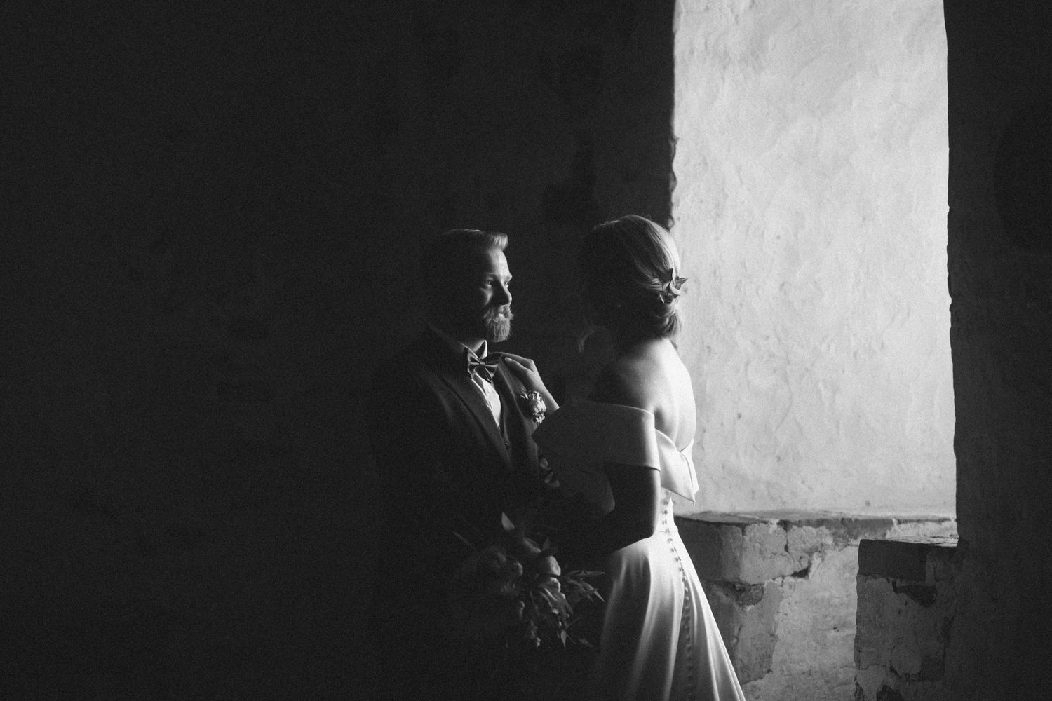 Black and white wedding portrait with couple standing by bright window in stone room.