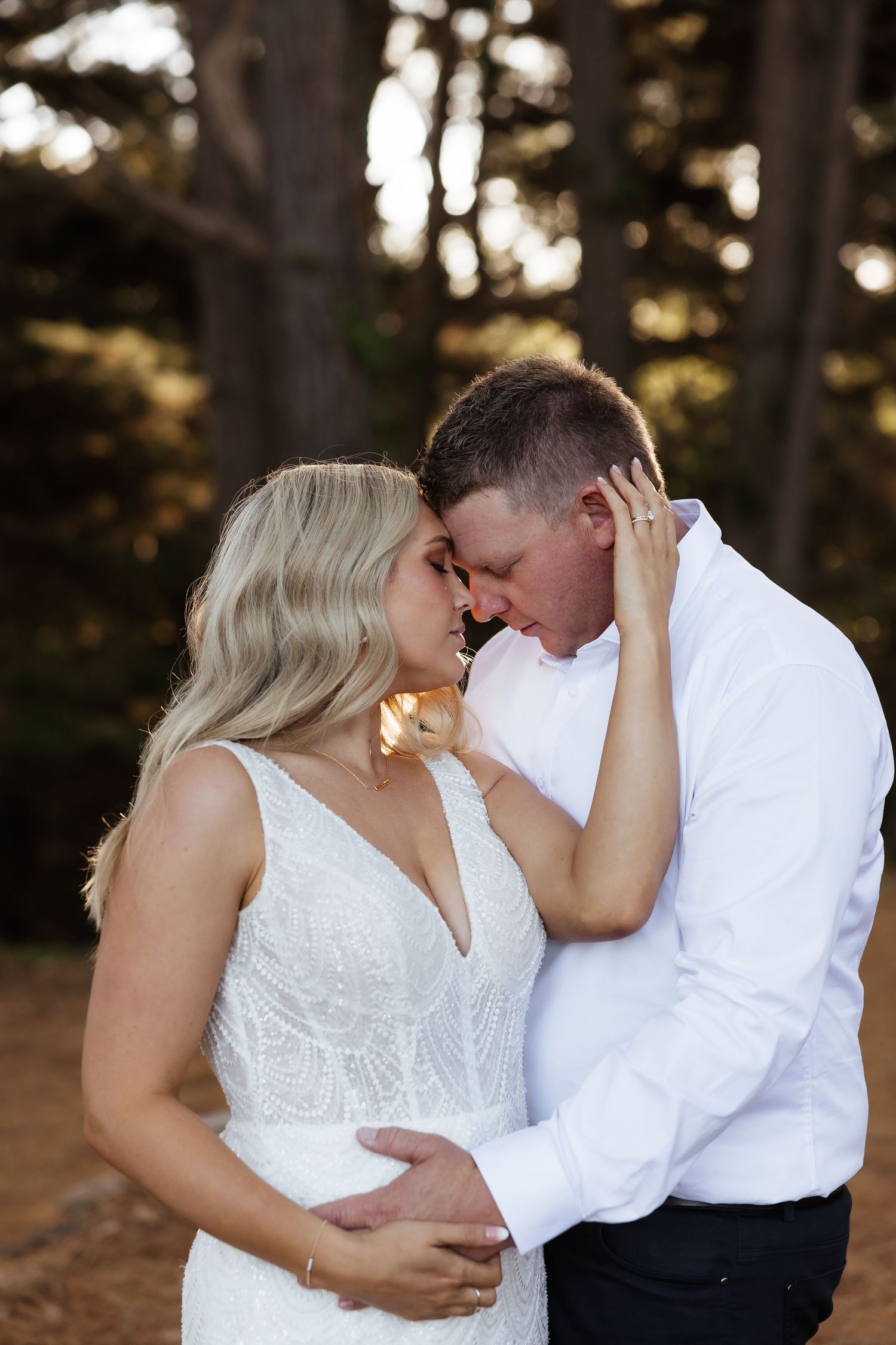 A romantic wedding couple share an intimate moment together in a forest setting at sunset.
