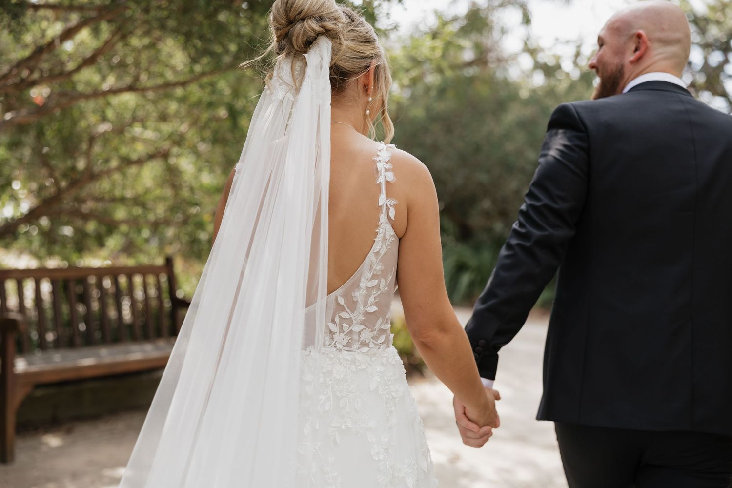 Bride and groom share a tender moment walking hand in hand through a sunlit forest path.