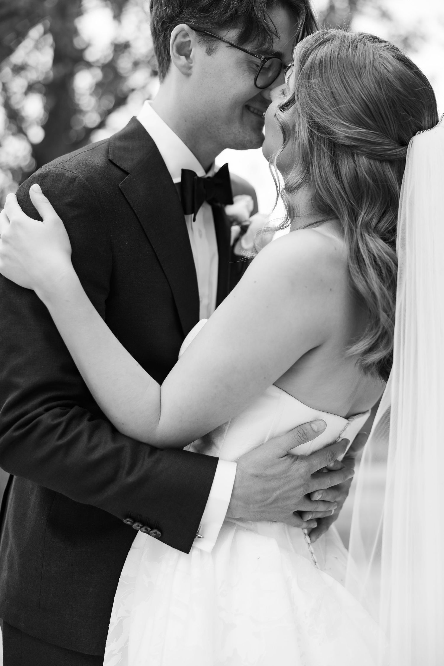 Black and white photo of a smiling groom in a tuxedo embracing his bride in a wedding dress and veil outdoors.